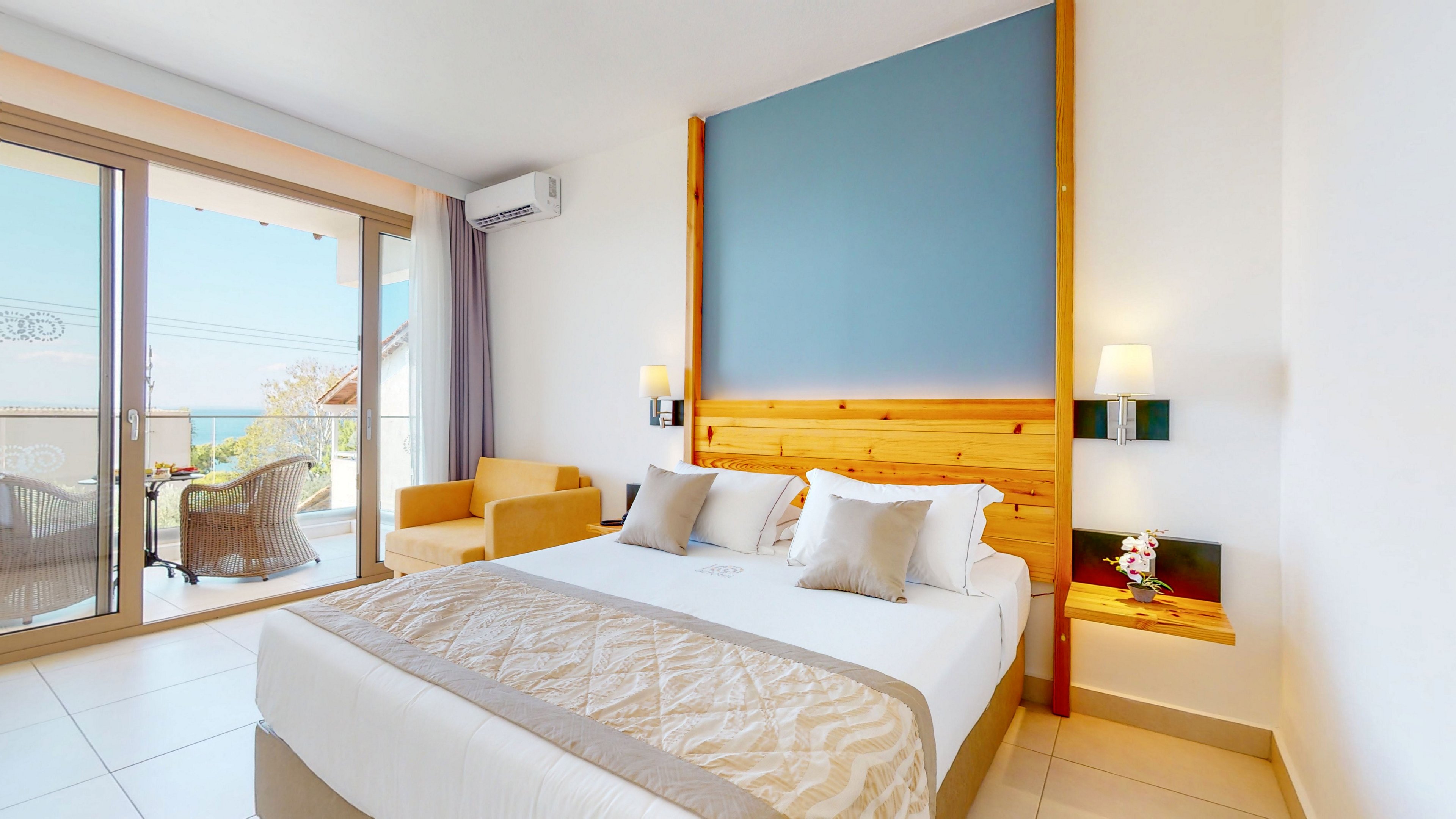 A contemporary hotel bedroom features a neatly made double bed with neutral bedding and accent pillows. The room includes a wooden headboard, wall-mounted lamps, and a small side table with decorative flowers. Large sliding glass doors open to a furnished balcony with wicker chairs and a table, offering an outdoor seating area and distant sea view. Soft natural light and a clean, minimalist design create a calm and inviting atmosphere.