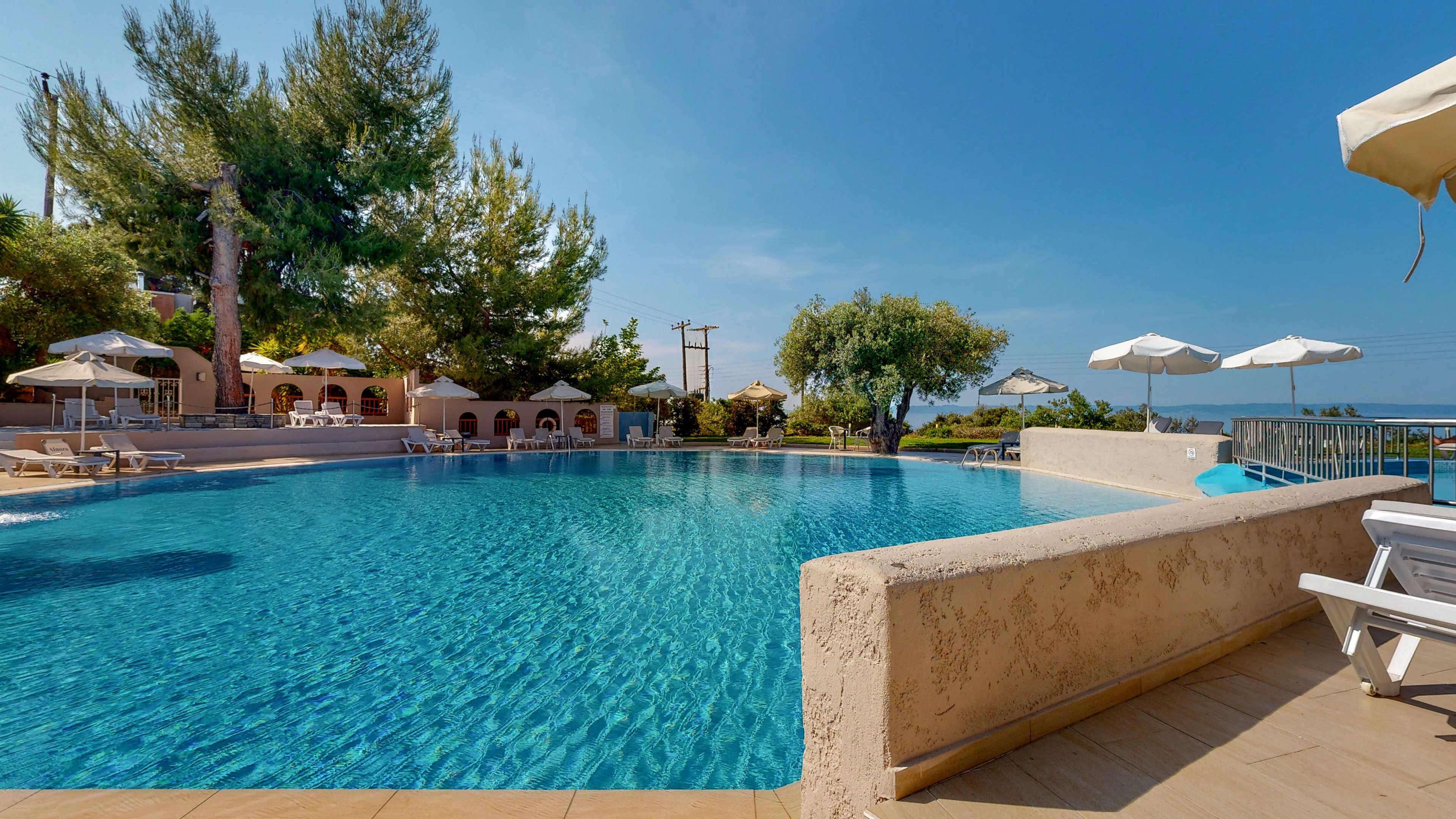 A large outdoor swimming pool glistens under a clear blue sky at a resort. Sun loungers and white umbrellas line the poolside terrace, offering shaded relaxation areas. Trees and low buildings frame the background, creating a tranquil holiday atmosphere. The scene suggests a peaceful, sunny day by the water with views extending toward the sea.
