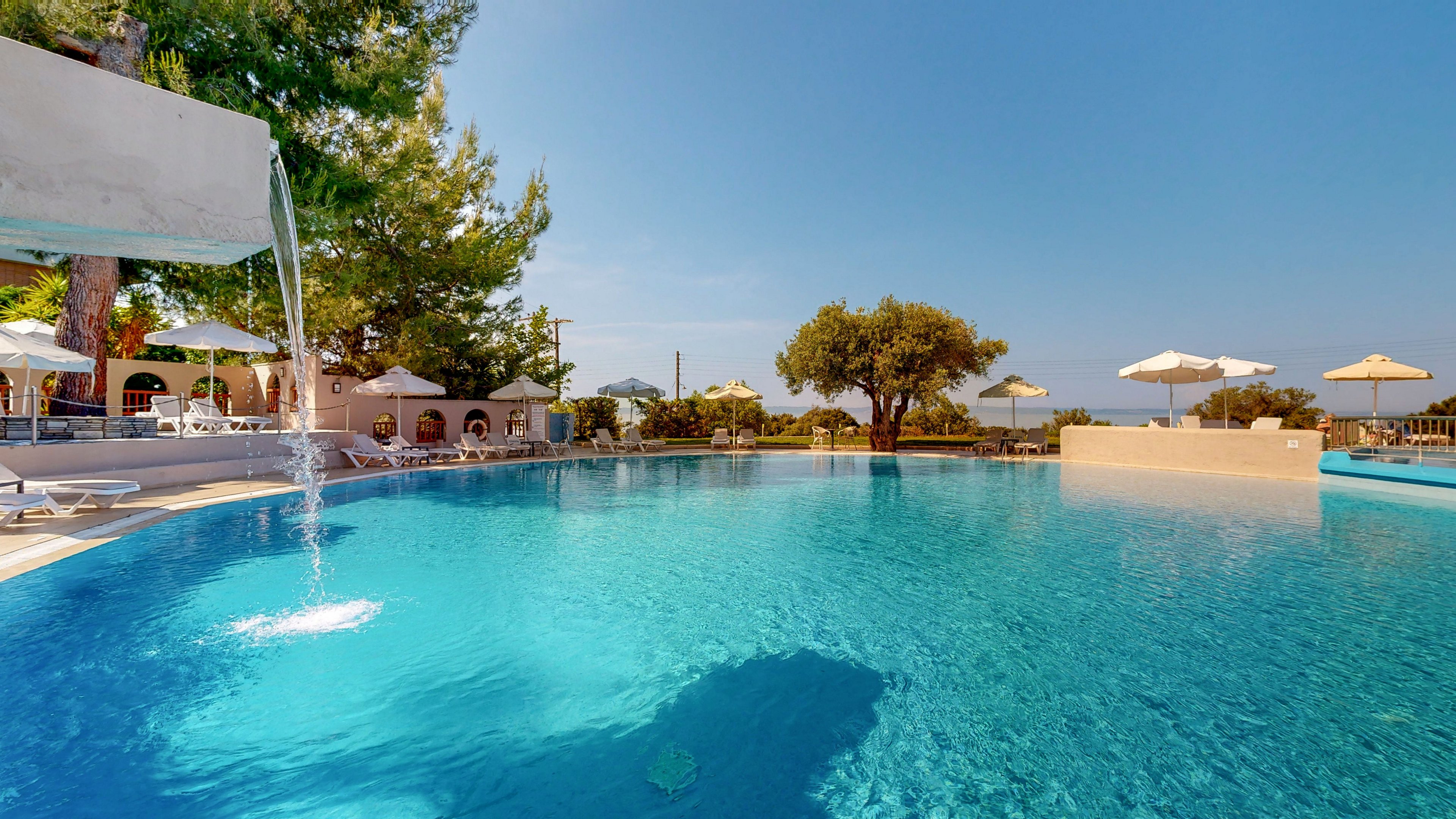 A large outdoor swimming pool glistens under a clear blue sky at a resort. Water cascades from a fountain feature into the pool, creating gentle ripples. Sun loungers with white umbrellas line the far side, set among trees and low buildings. The scene conveys a relaxed vacation atmosphere in a bright, warm climate.