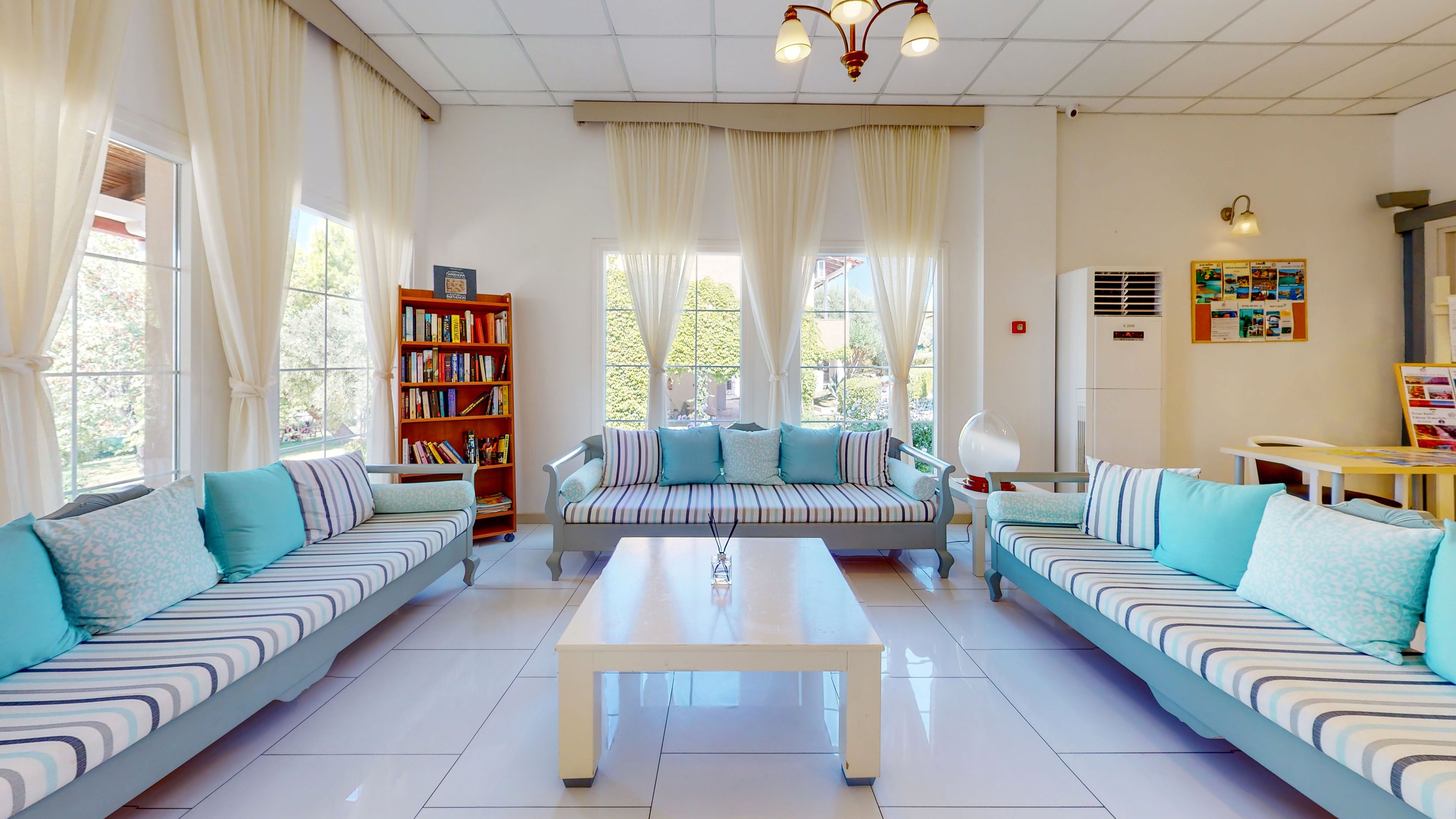 A spacious living room features long striped sofas with light blue and white cushions arranged around a simple white coffee table. Large windows with sheer curtains let in abundant natural light and reveal greenery outside. A wooden bookcase filled with books stands to the left, while a wall-mounted air conditioner and colorful wall art appear on the right. The overall palette is airy and coastal, dominated by whites, soft blues, and natural wood accents.