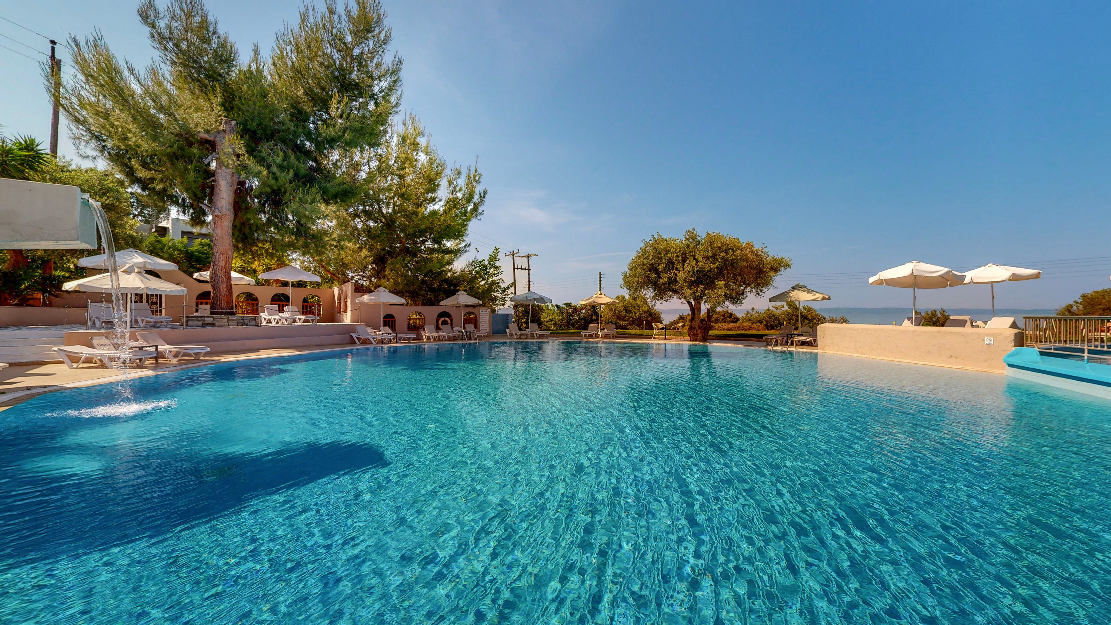 A large outdoor swimming pool glistens under clear blue skies at a resort. White sun loungers and umbrellas line the poolside terrace, offering shaded relaxation areas. Tall trees and greenery surround the scene, adding a natural backdrop to the tranquil water. The overall atmosphere is calm and inviting, suggesting a warm vacation destination.