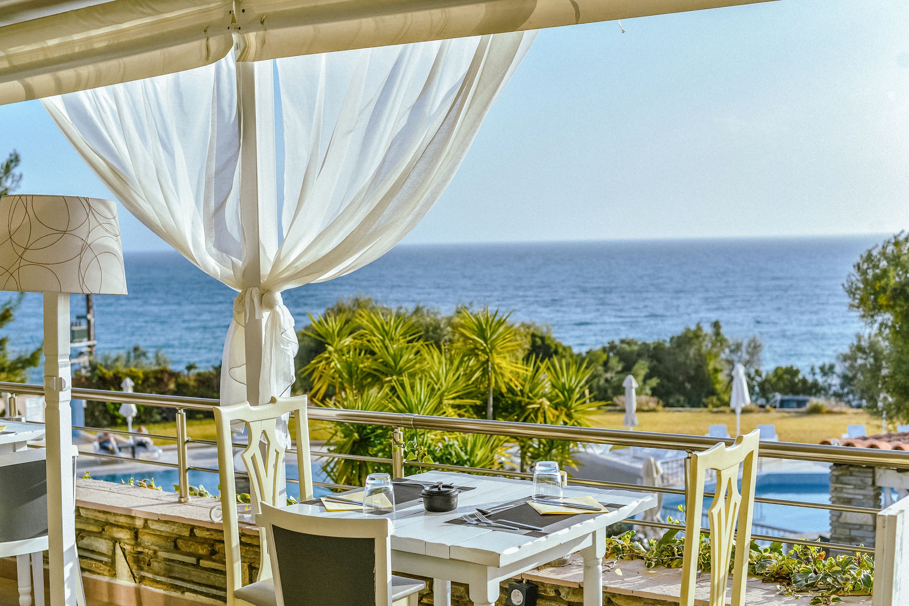 An elegant outdoor restaurant terrace overlooks a calm blue ocean. White curtains flow gently around neatly set tables with glassware and table settings. In the background, a swimming pool, sun loungers, and lush palm-like plants create a resort atmosphere. The bright daylight and soft colors convey a relaxed, vacation mood.