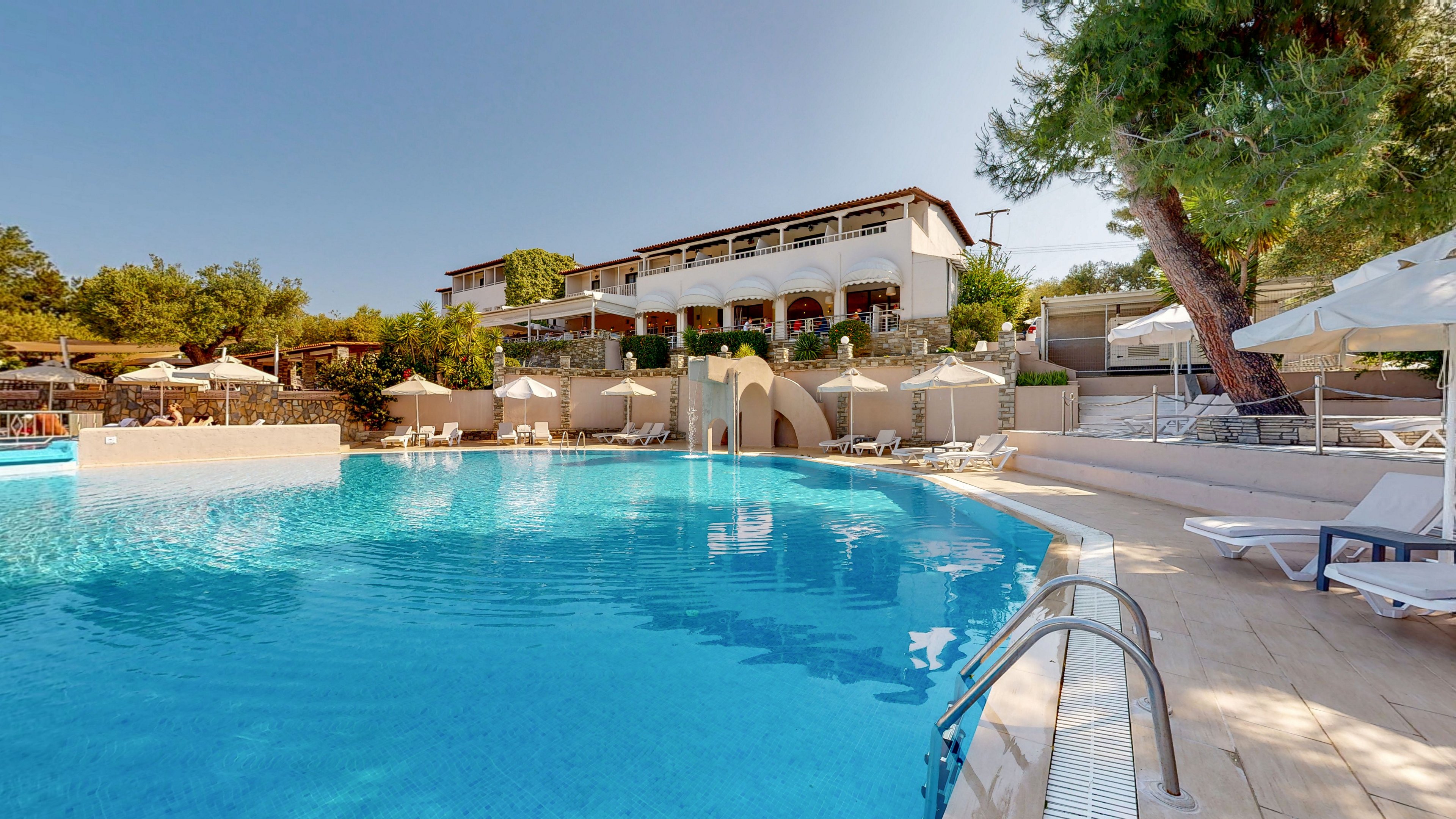 A large outdoor swimming pool is shown in front of a multi-story resort building. Sun loungers and umbrellas line the poolside terrace, creating a relaxing vacation atmosphere. Clear blue water reflects the bright sky, while trees and greenery surround the property. The scene suggests a warm, sunny day at a holiday hotel or resort.