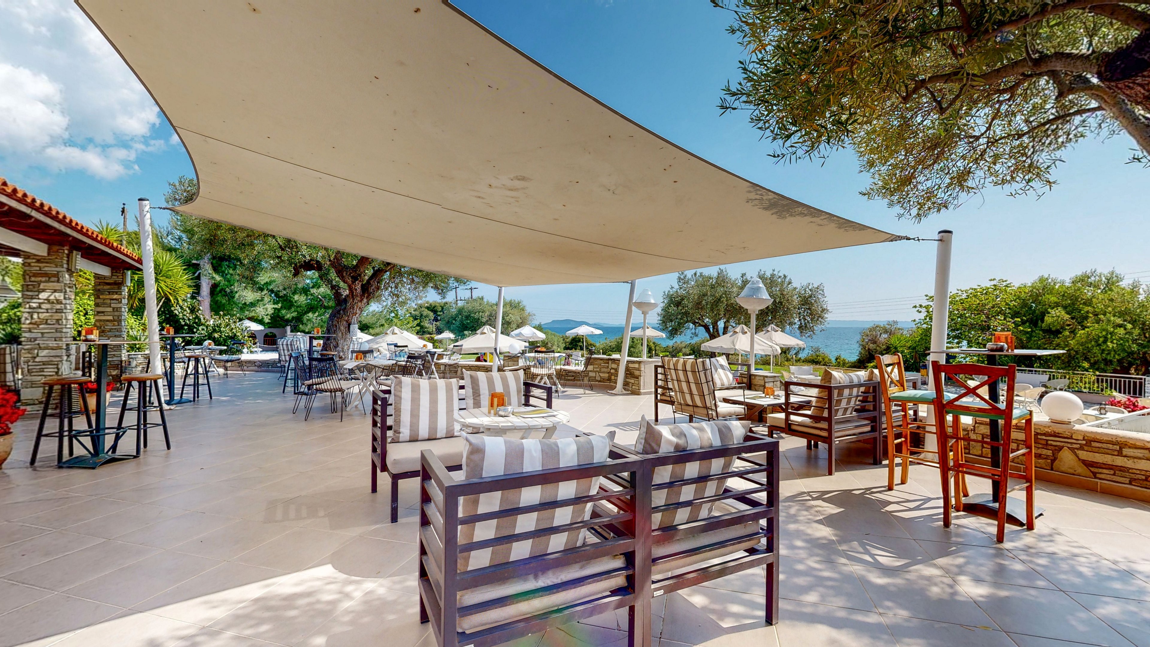 An open-air terrace cafe features striped cushioned chairs and wooden tables under a large sail shade. The setting overlooks a scenic coastline with blue water visible in the distance. Umbrellas, greenery, and mature trees surround the seating area, creating a relaxed holiday atmosphere. The bright daylight and clean tiled floor emphasize a casual, inviting Mediterranean-style outdoor space.