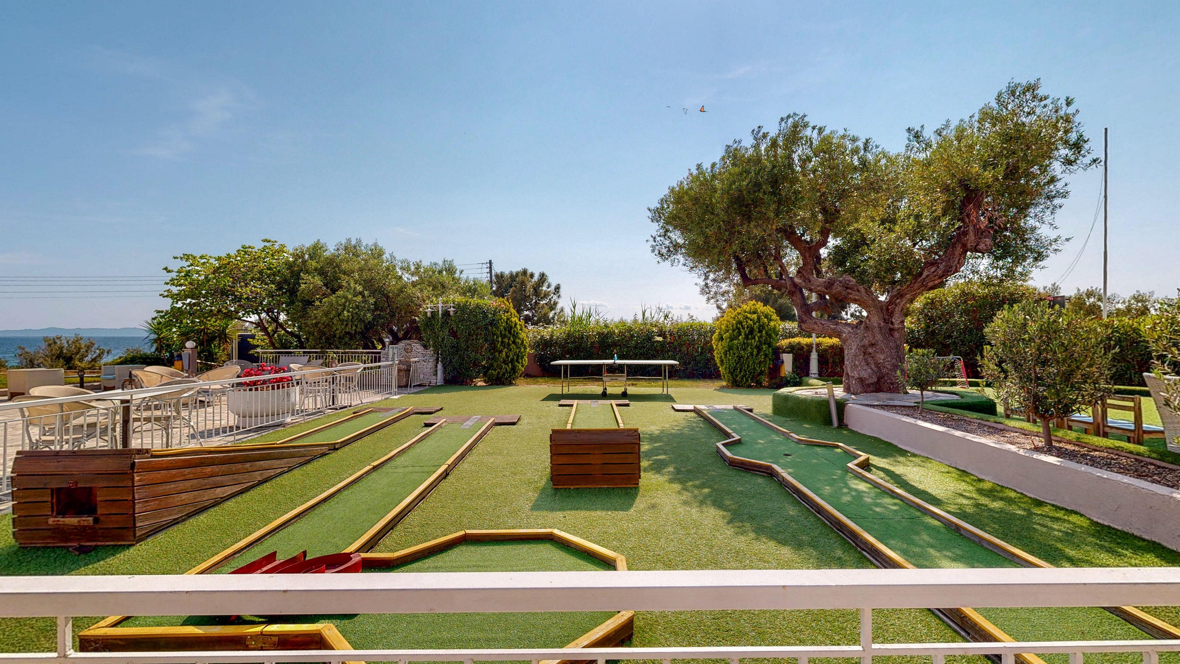 An outdoor mini golf course is set on a neatly maintained artificial grass terrace. Multiple wooden-bordered lanes and obstacles create a playful layout under a clear blue sky. Mature trees and trimmed shrubs frame the area, with seating and railings along the side. In the distance, the sea is visible, adding a scenic coastal backdrop to the recreational setting.