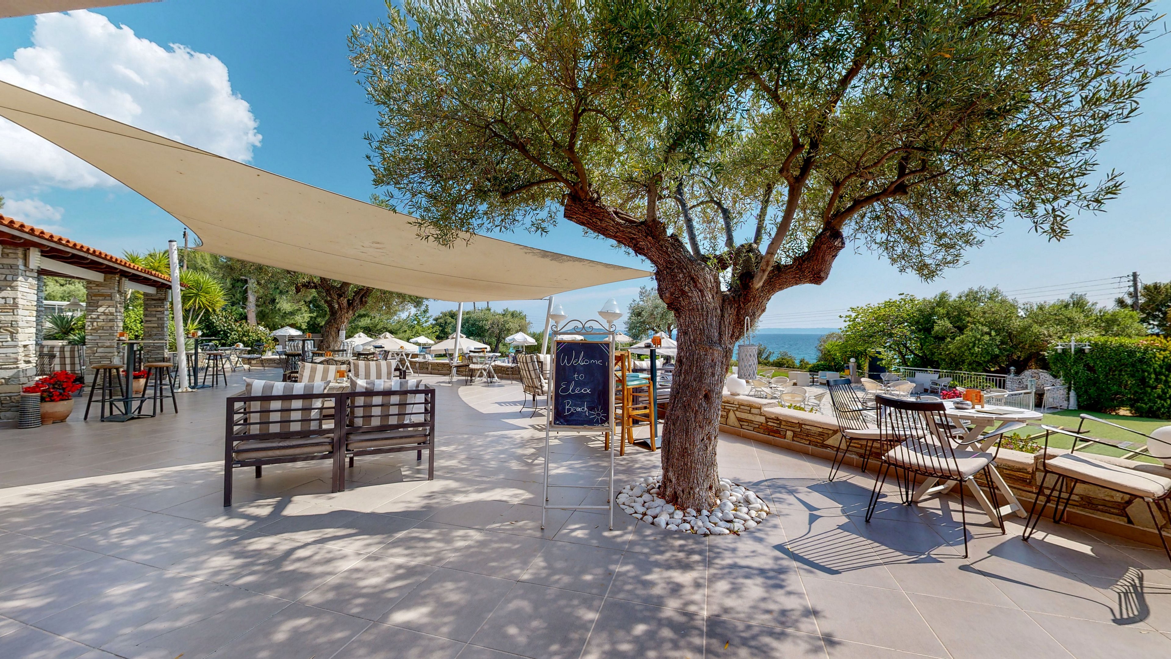 An inviting outdoor café terrace is set beneath a large tree, with tables and chairs arranged for guests. A sail-like canopy provides shade over part of the tiled patio, while sun loungers and umbrellas are visible near a pool or garden area in the background. A chalkboard menu stands in the center, adding a casual bistro feel to the scene. The bright daylight, blue sky, and distant sea create a relaxed Mediterranean atmosphere.