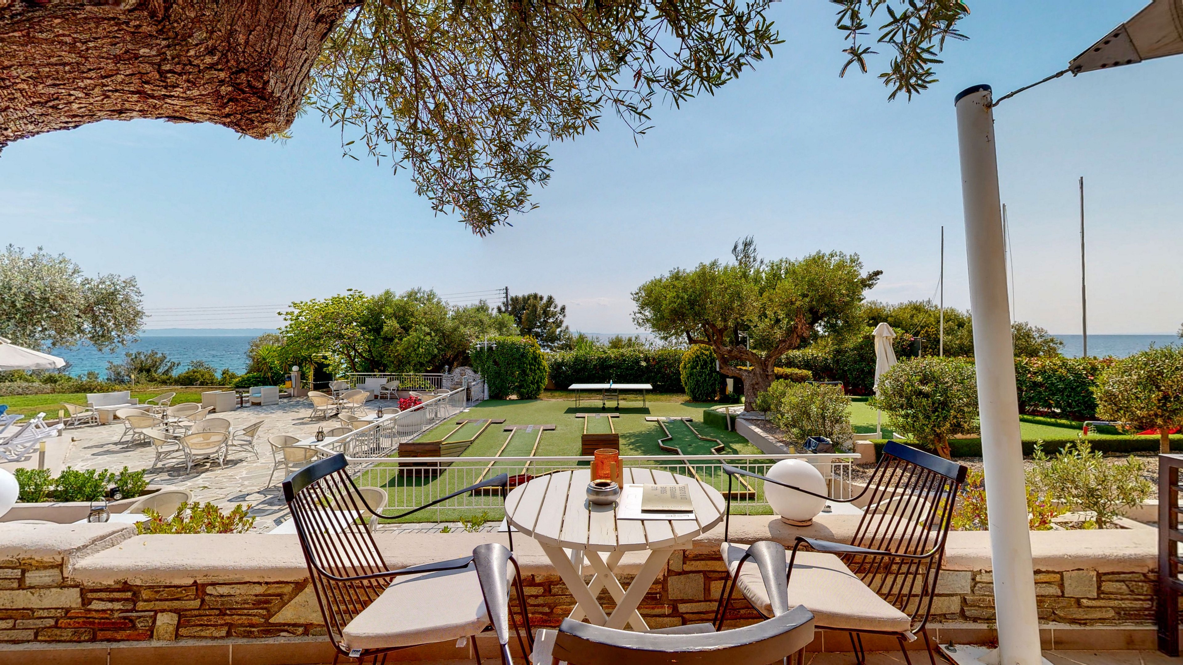 A cozy terrace with two chairs and a small round table overlooks a landscaped garden and the sea in the distance. The foreground features a shaded seating area with a tree, stone wall, and decorative lantern on the table. Beyond the terrace, manicured lawns, hedges, and outdoor furniture create a relaxed resort atmosphere. The calm blue water and clear sky suggest a sunny, peaceful coastal day.