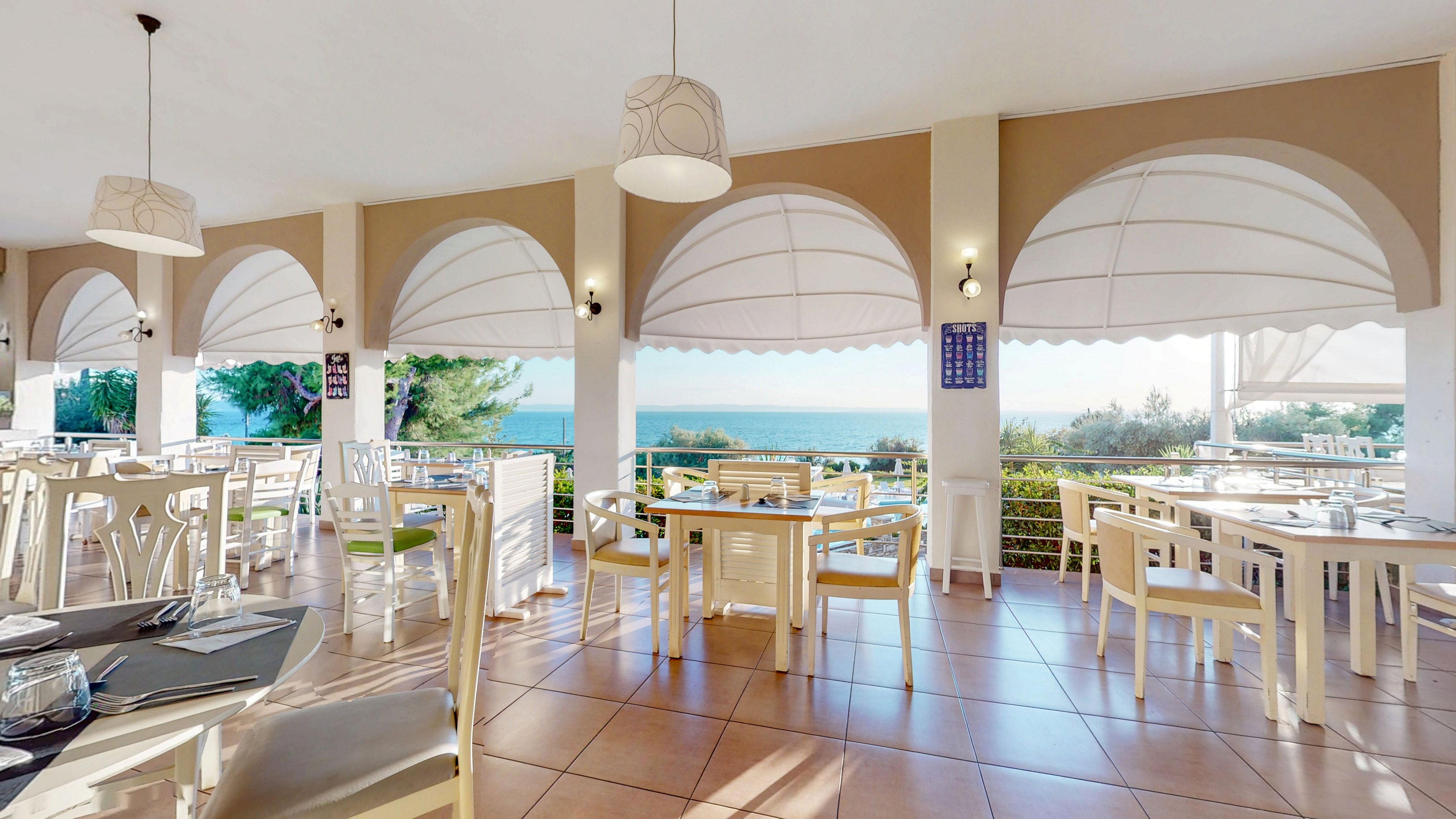 An airy, open-sided restaurant overlooks a calm blue sea. White tables and chairs are neatly arranged on a tiled floor under large arched openings with fabric awnings. Sunlight streams through the arches, casting soft shadows and enhancing the coastal atmosphere. The space feels relaxed and inviting, ideal for casual dining with scenic waterfront views.