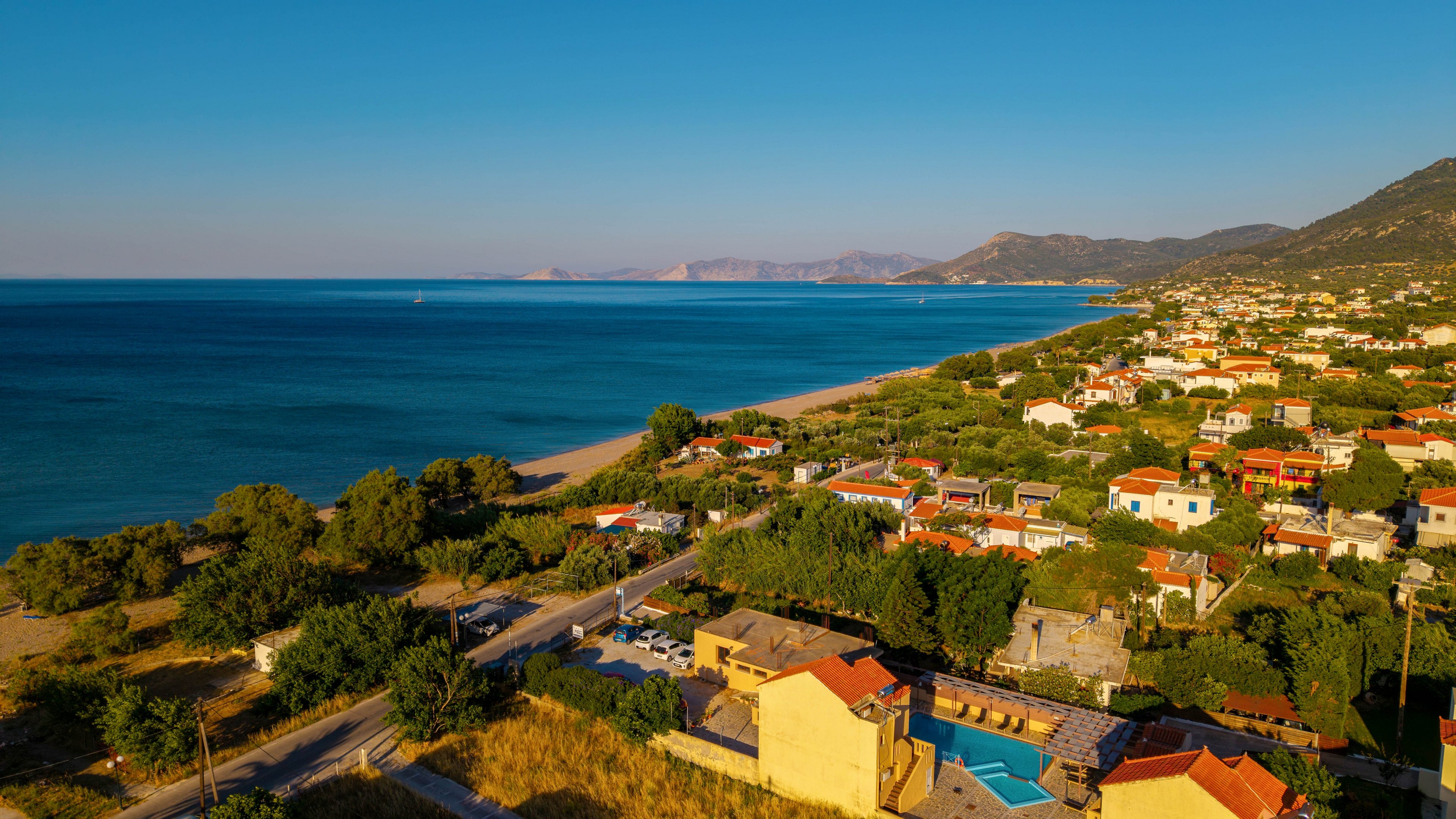 An aerial view captures a coastal village stretching along a calm blue shoreline. Colorful houses with red and yellow roofs are nestled among green trees and vegetation. A narrow road runs parallel to the beach, connecting the homes and small buildings. The distant hills and mountains frame the horizon under a clear blue sky, suggesting a warm, tranquil evening. The overall scene conveys a peaceful seaside community bathed in golden light.