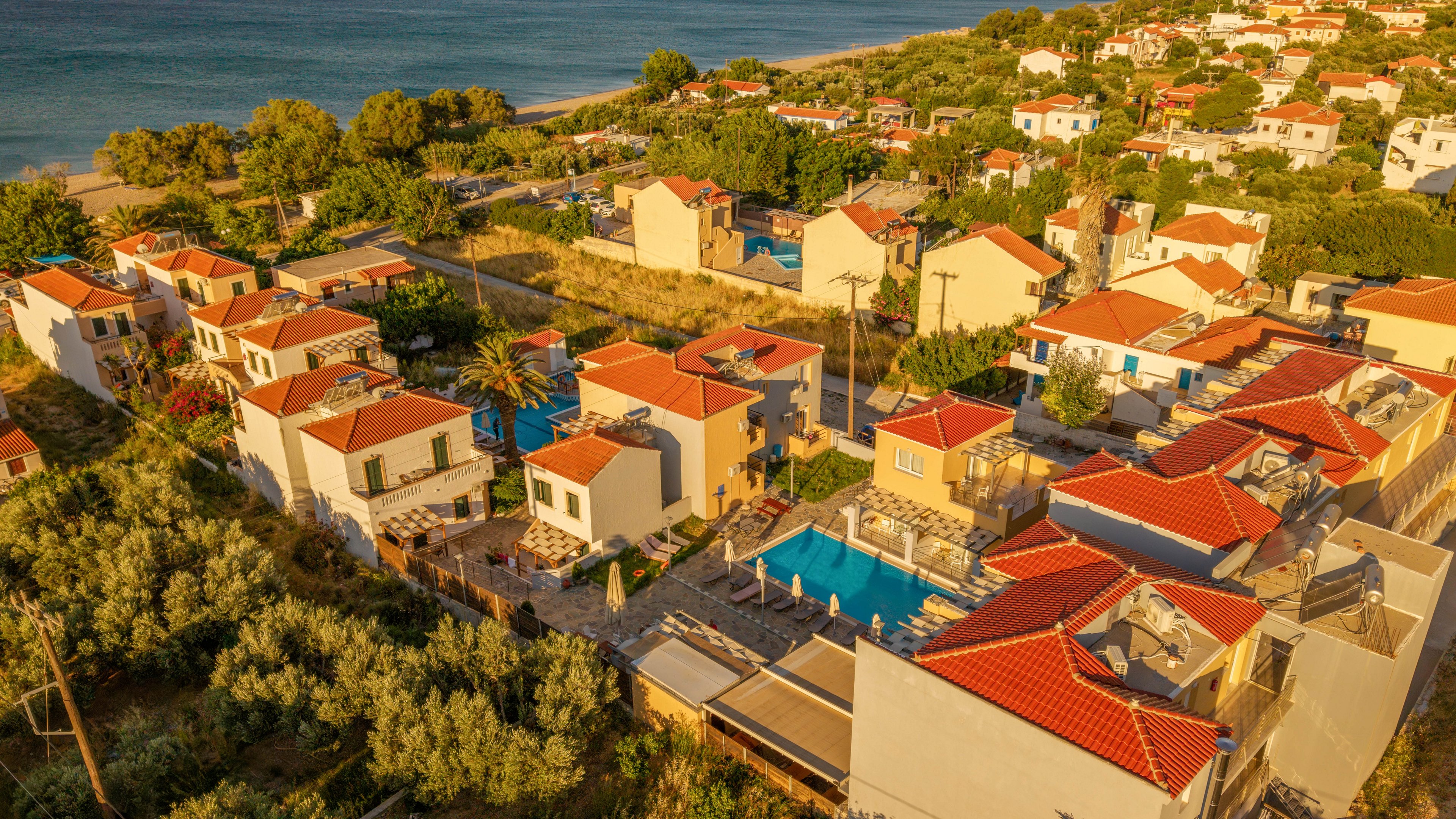 An aerial view shows a coastal village with clusters of white houses topped with red tile roofs. The neighborhood is surrounded by lush greenery and trees, with the sea visible in the background. A central swimming pool and sunlit courtyards suggest a relaxed vacation setting. Warm golden hour light enhances the tranquil, Mediterranean-style atmosphere.