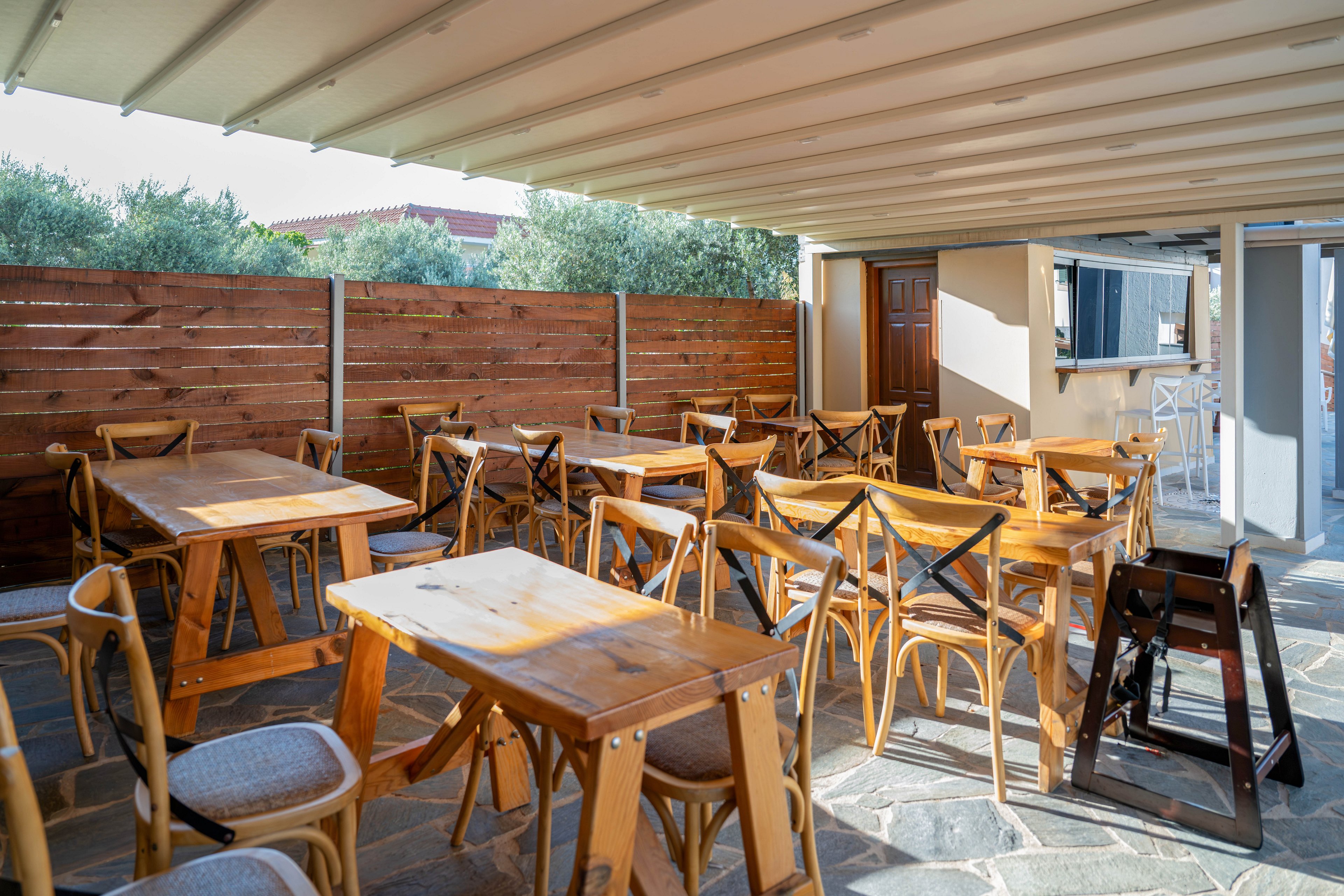 An empty outdoor cafe terrace features multiple wooden tables and chairs neatly arranged under a covered pergola. Sunlight streams in from the open side, casting soft shadows on the stone floor. A wooden privacy fence lines the perimeter, with greenery and a building visible beyond it. The scene feels calm and inviting, suggesting a quiet moment before guests arrive.