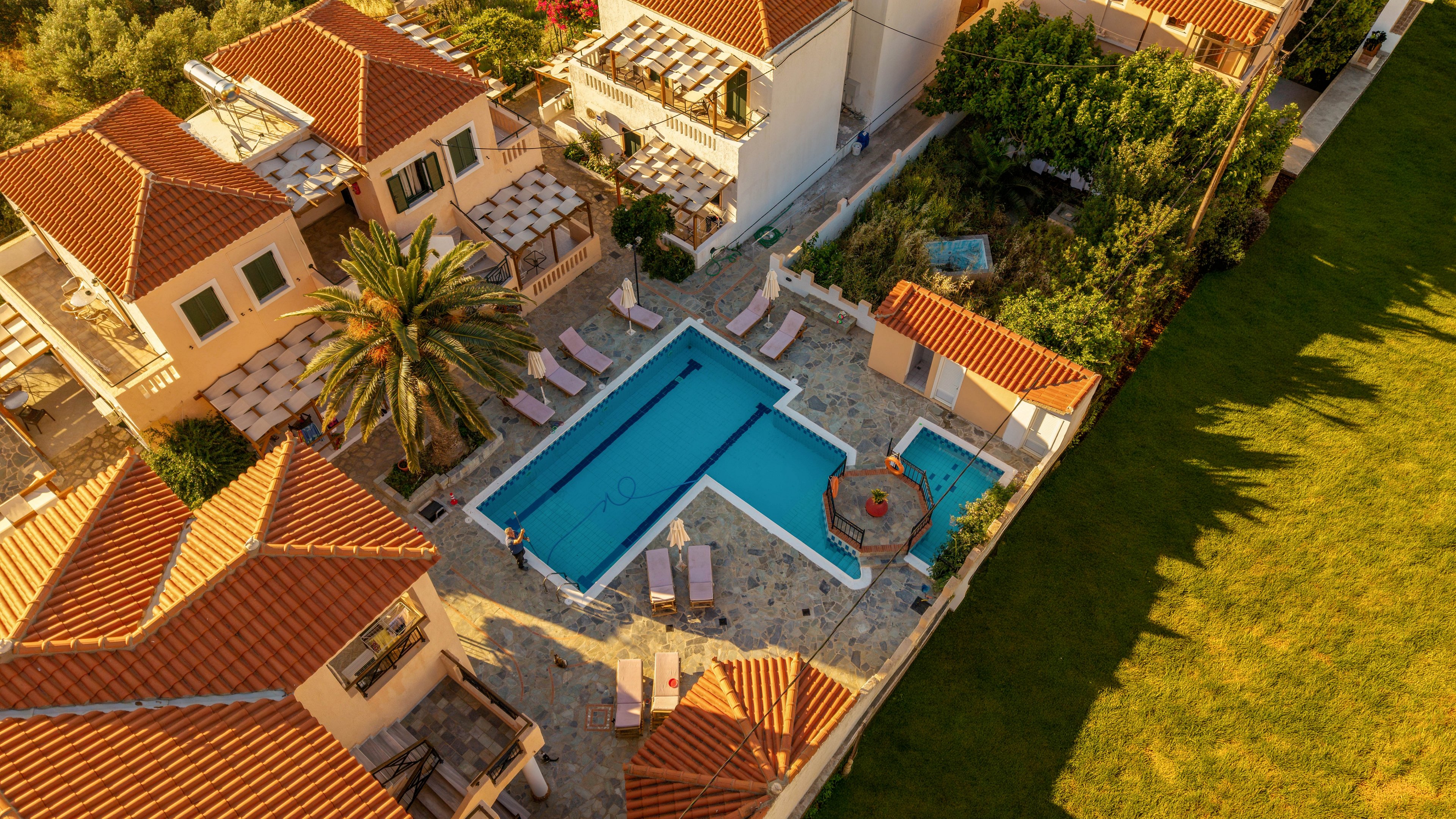 An aerial view shows a rectangular swimming pool and adjacent hot tub surrounded by a stone patio. Several sun loungers and umbrellas are arranged neatly around the pool area, suggesting a relaxing resort or villa setting. Terracotta-roofed buildings and a tall palm tree frame the courtyard, while a large green lawn borders one side. Warm sunlight casts long shadows, enhancing the tranquil, vacation-like atmosphere.