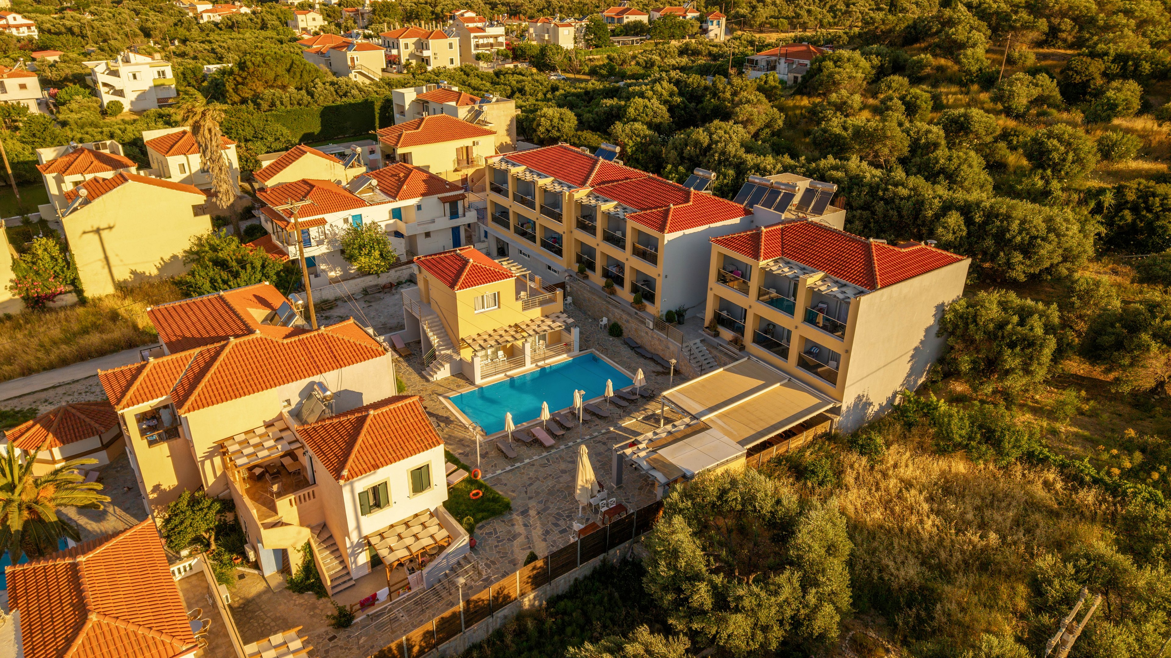 An aerial view shows a Mediterranean-style hotel complex with multiple cream-colored buildings and red-tiled roofs. A central swimming pool with sun loungers and umbrellas forms the focal point of the courtyard. The property is surrounded by lush greenery, trees, and neighboring houses in a hilly landscape. Warm golden-hour sunlight enhances the colors and creates a relaxed vacation atmosphere.