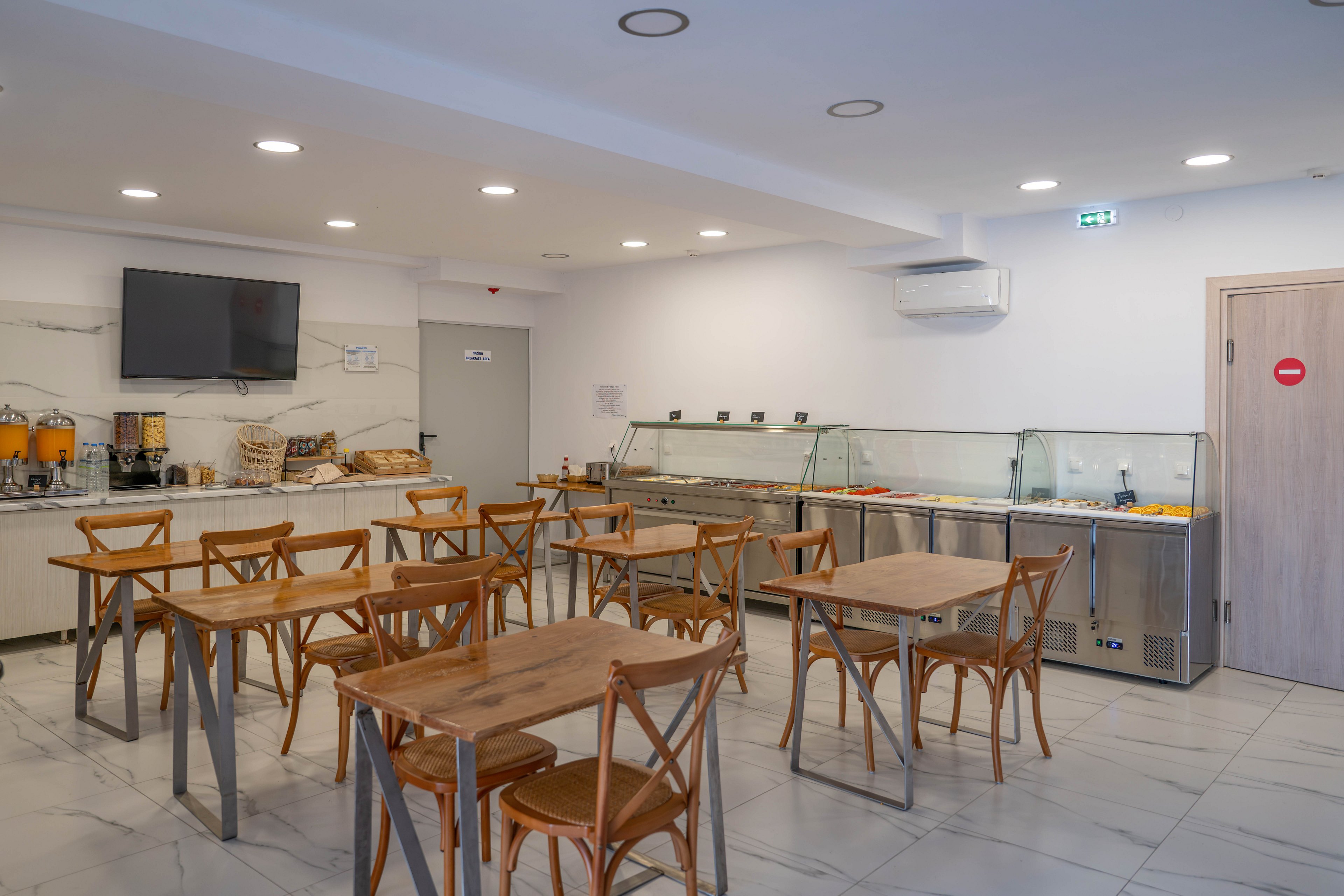 A clean, modern breakfast room features several wooden tables and chairs arranged neatly on a tiled floor. Along the back wall, a self-service buffet area includes juice dispensers, bread, and covered food trays in refrigerated counters. A large flat-screen television is mounted above the counter, and an air conditioning unit is visible on the wall. Bright ceiling lights and neutral decor create a calm, minimalist atmosphere.