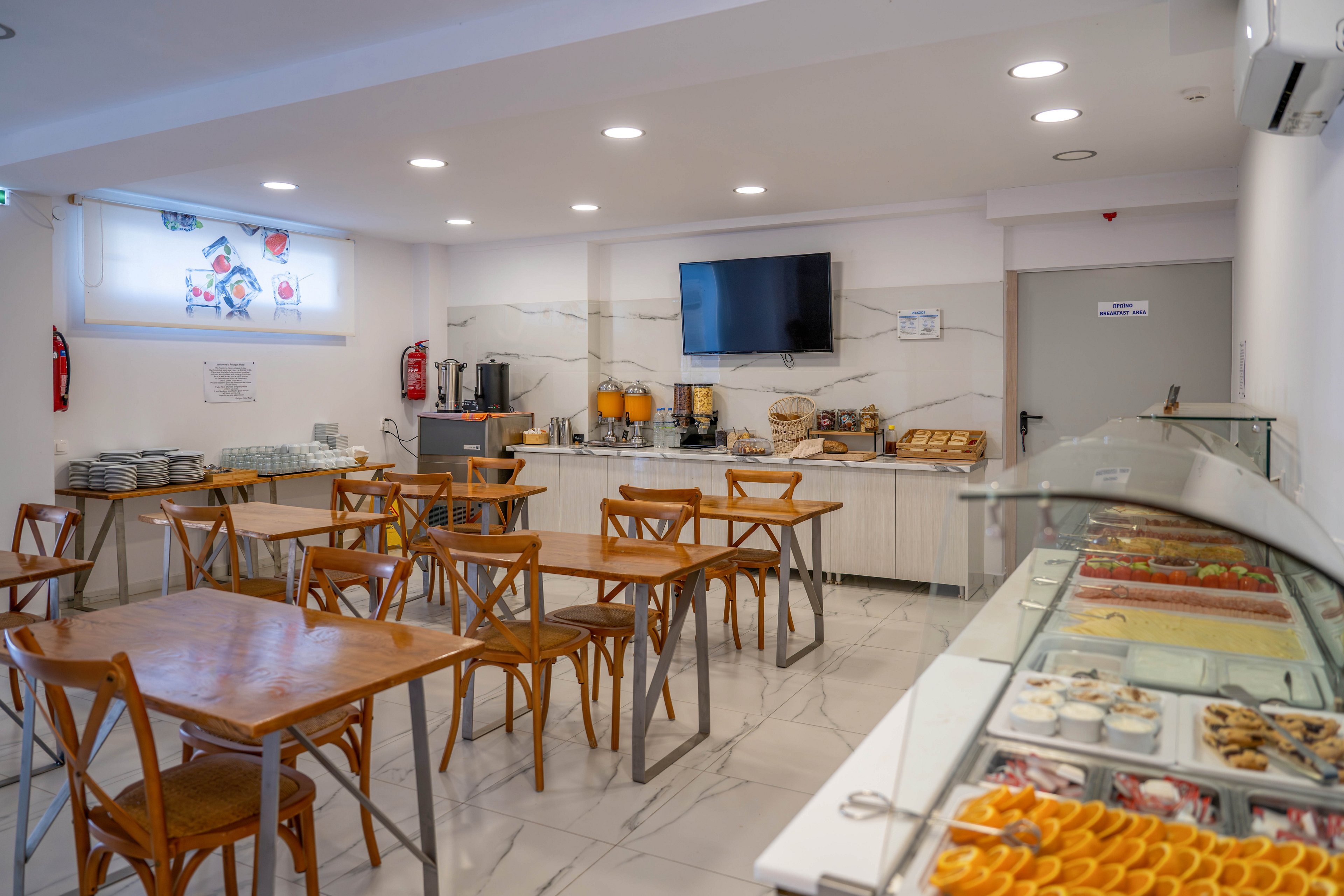 A clean, modern breakfast room features several wooden tables and chairs arranged neatly on a polished floor. Along the back wall, a self-service buffet offers beverages, bread, and cereals beneath a mounted flat-screen television. On the right, a glass display case showcases assorted pastries and desserts, including orange slices and small cakes. Recessed ceiling lights and a large window provide bright, even illumination, creating a welcoming dining atmosphere.