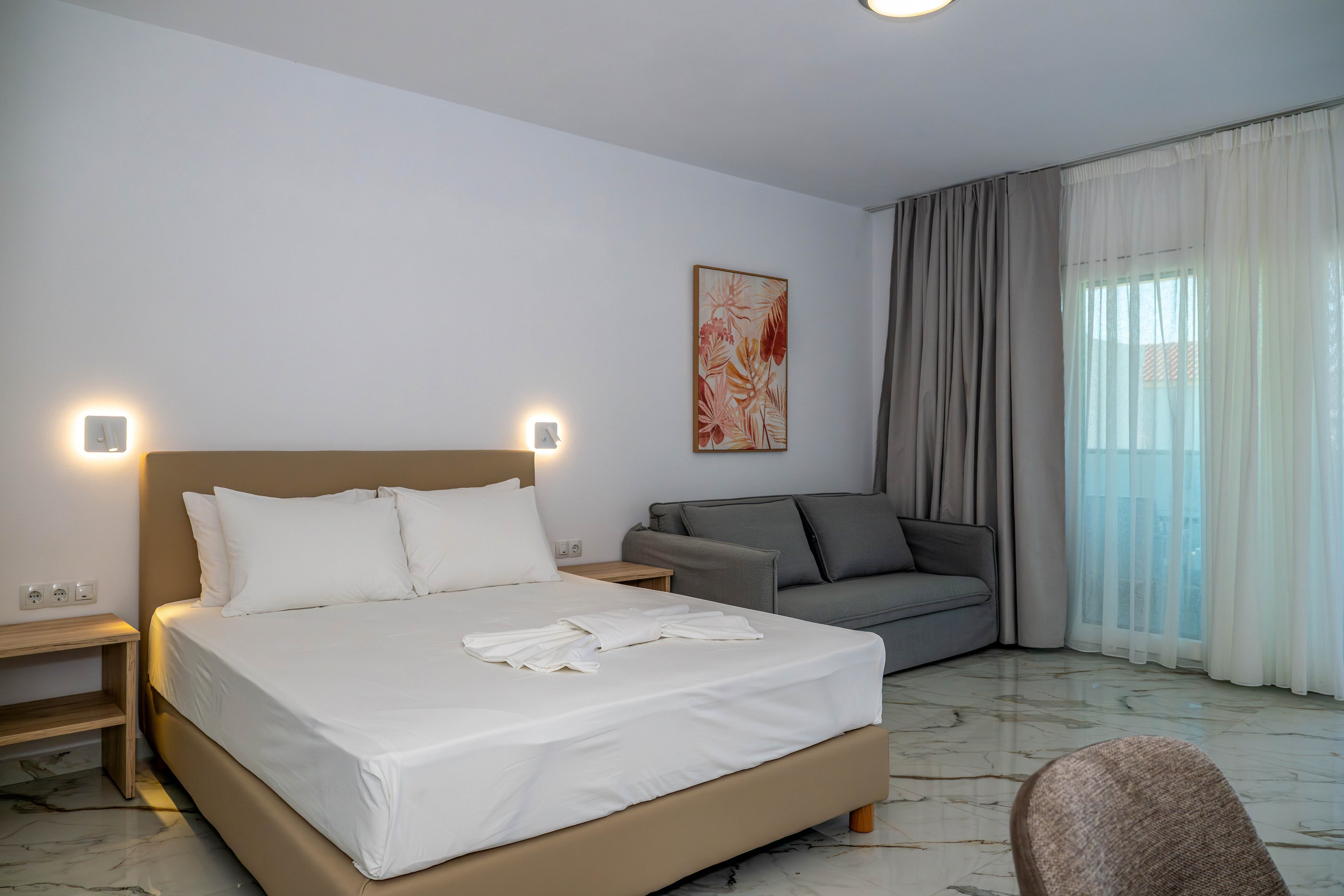 A contemporary hotel bedroom features a double bed with white linens and pillows on a beige frame. A gray sofa sits against the wall beside floor-to-ceiling windows covered with sheer and blackout curtains. Wall-mounted lights flank the bed, and a framed abstract artwork adds a decorative touch. Marble-style flooring and neutral tones create a clean, minimalist atmosphere.