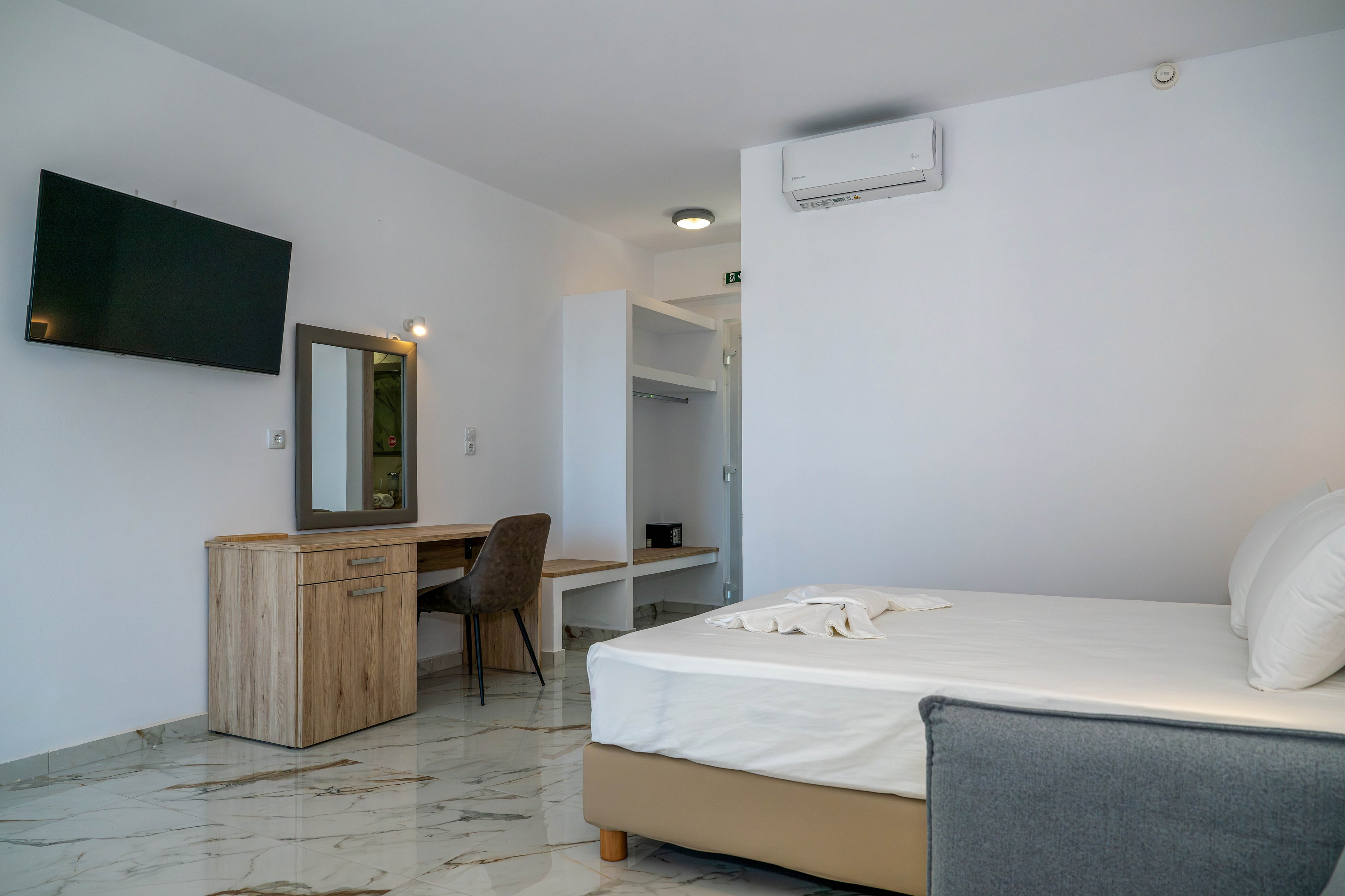 A contemporary hotel room features a neatly made bed with white linens and a gray upholstered frame. A wooden desk with a mirror and cushioned chair sits against the wall, illuminated by a small wall light. A flat-screen television is mounted on the opposite wall, and an open wardrobe area with shelves and a safe is visible in the background. The space has white walls, marble-style flooring, and a wall-mounted air conditioner, creating a clean and minimalist atmosphere.