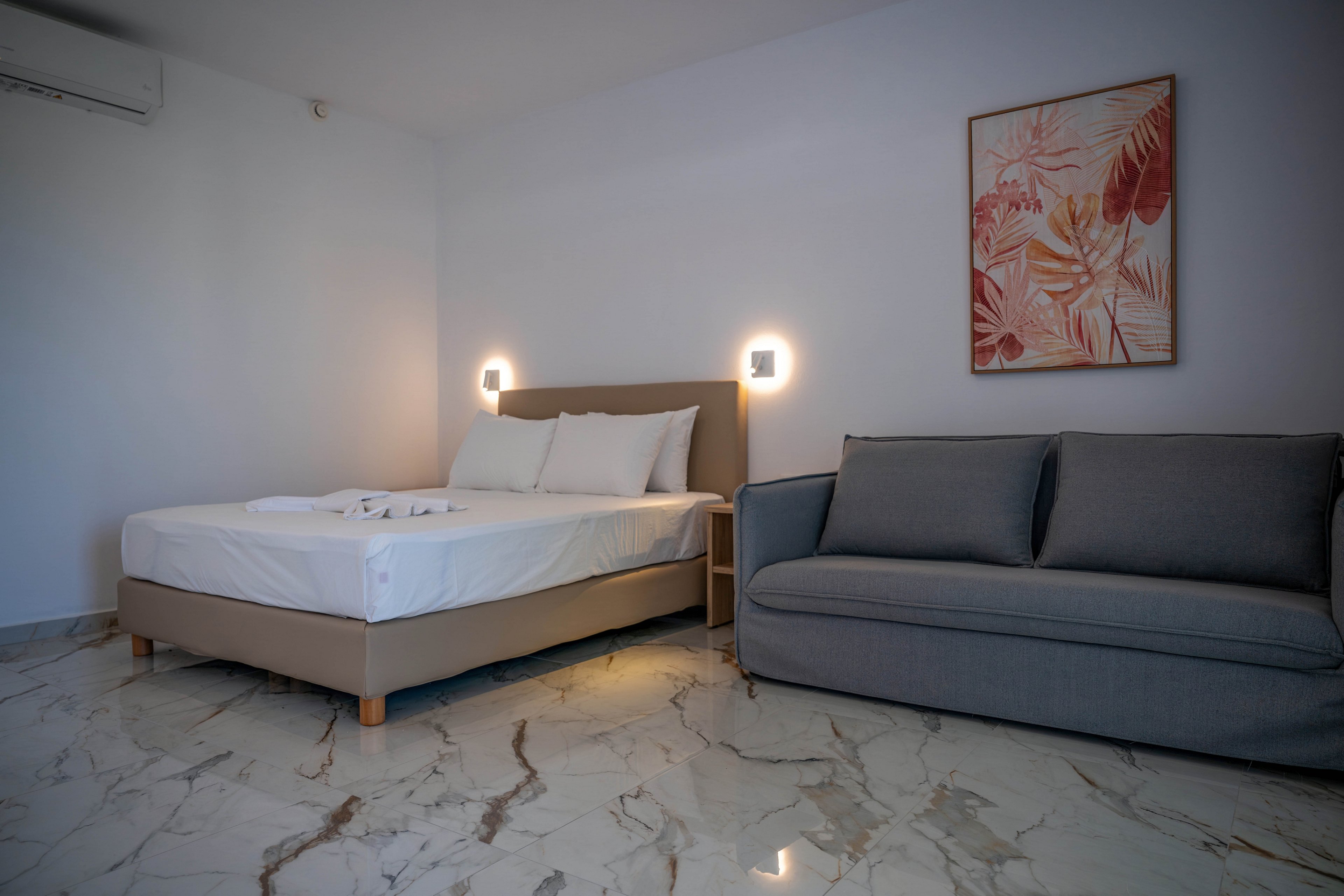 A modern minimalist bedroom features a double bed with white linens and folded towels placed on top. A gray upholstered sofa sits beside the bed, creating a small lounge area within the room. Two wall-mounted lights cast a warm glow on the white walls, complemented by a framed botanical artwork. The polished marble floor adds a sleek, contemporary feel to the interior space.