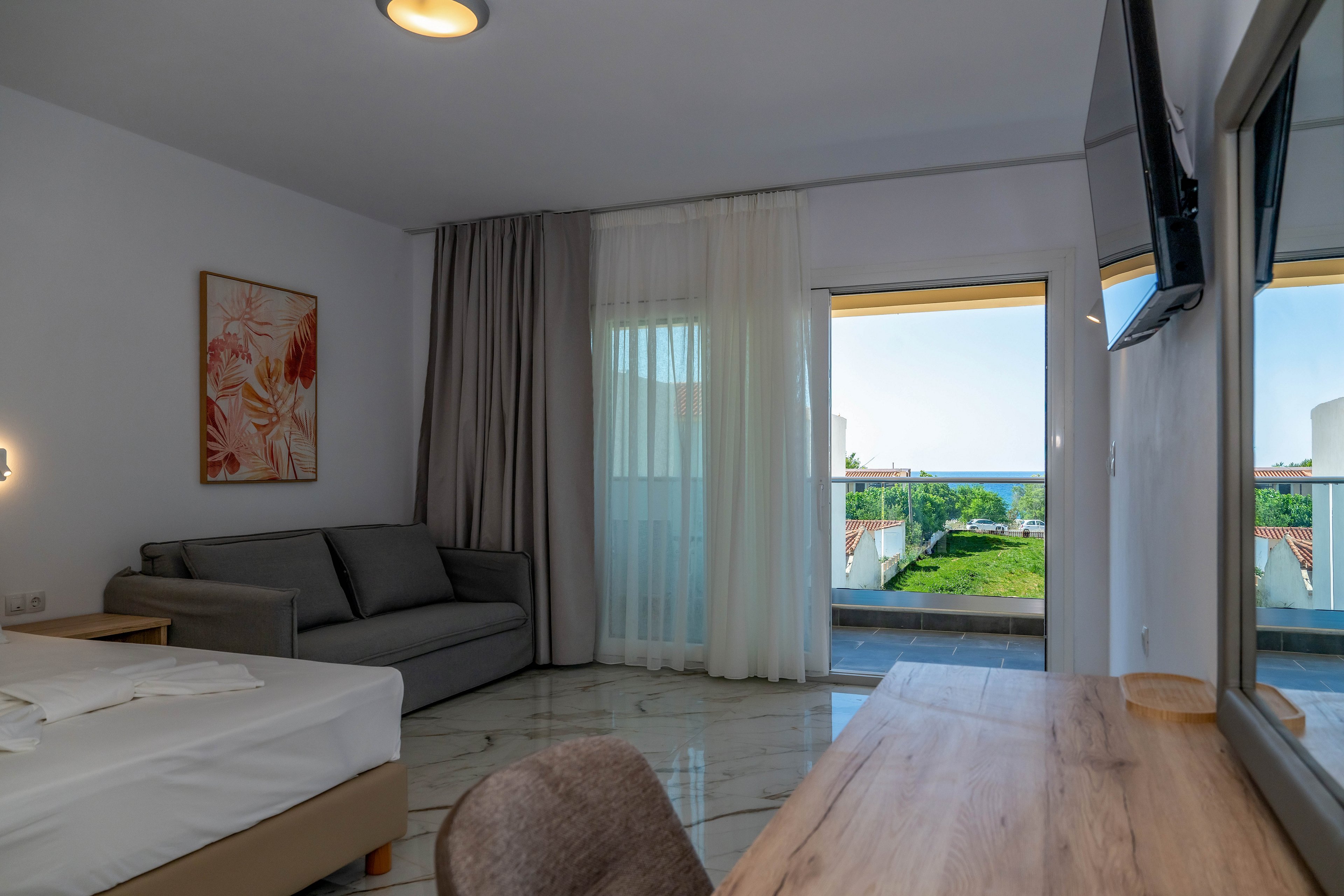 Contemporary hotel room interior featuring a bed, gray sofa, and wooden desk. Large sliding glass doors open to a balcony overlooking greenery and a distant sea view. Sheer white curtains and darker drapes frame the window, softening the natural light. A wall-mounted television and minimalist artwork complete the clean, modern aesthetic.