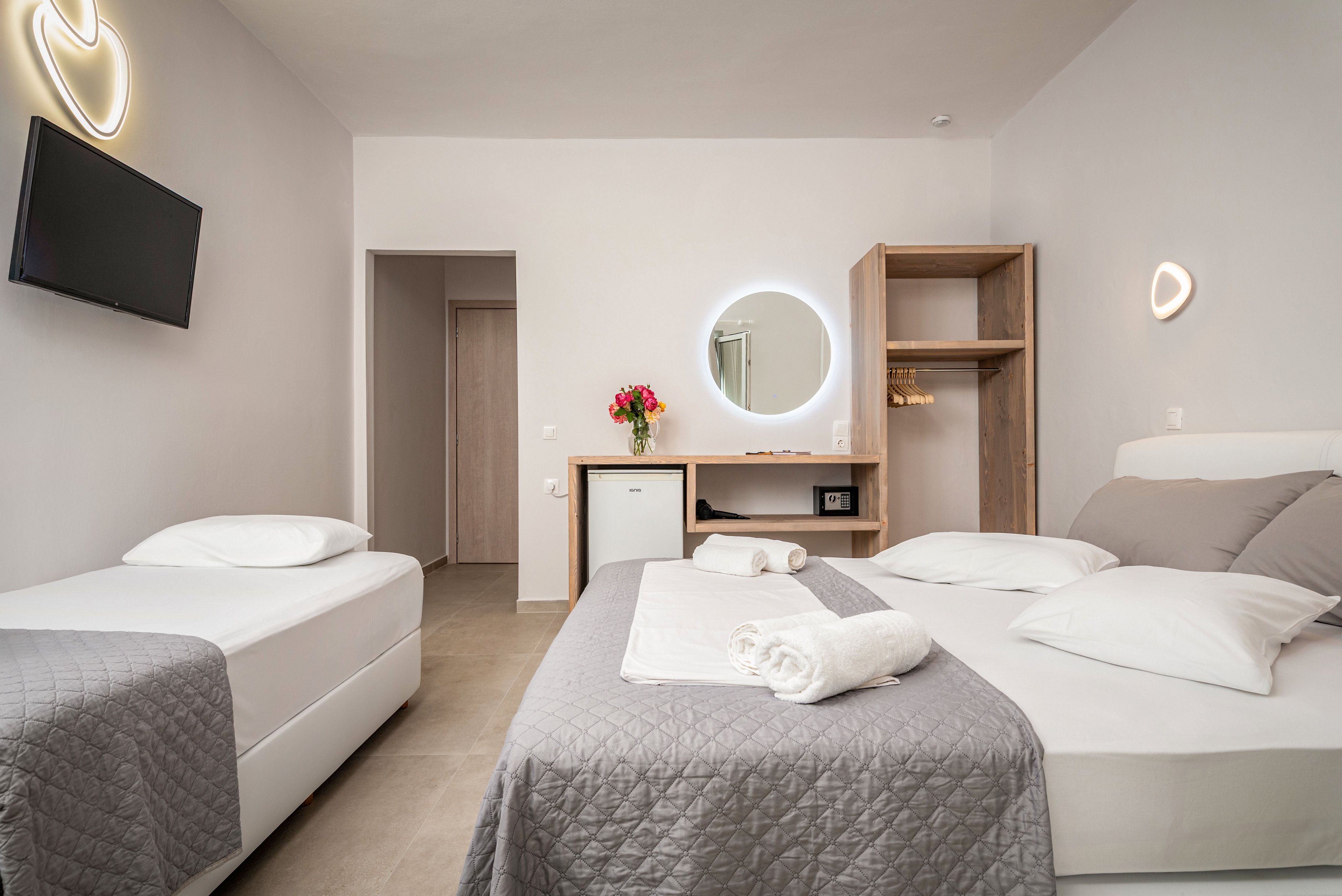 Contemporary hotel room interior featuring a double bed and a single bed with gray quilted covers and white pillows. The space includes a minimalist desk area with a round illuminated mirror, open shelving, and a small safe. Soft wall lighting, a wall-mounted television, and neutral-toned walls create a calm, inviting atmosphere. Rolled white towels on the main bed and a vase of flowers add a welcoming touch.