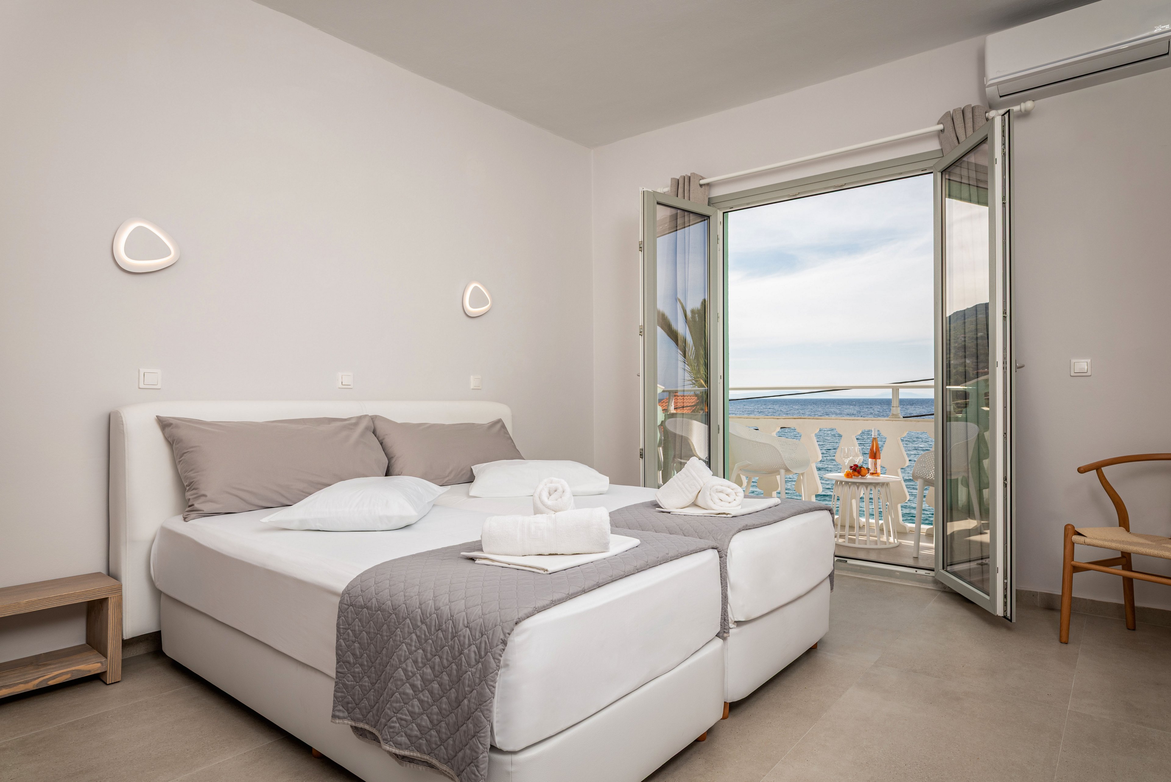 Modern double bedroom with neatly made twin beds and folded white towels is shown in a bright, minimalist interior. Neutral gray and white bedding complements the light walls and tiled floor, creating a calm and clean atmosphere. Glass doors open to a balcony with chairs and a table overlooking the sea and blue sky, suggesting a relaxing coastal vacation setting. Simple wall lights, a wooden chair, and an air conditioning unit add comfort and functionality to the space.