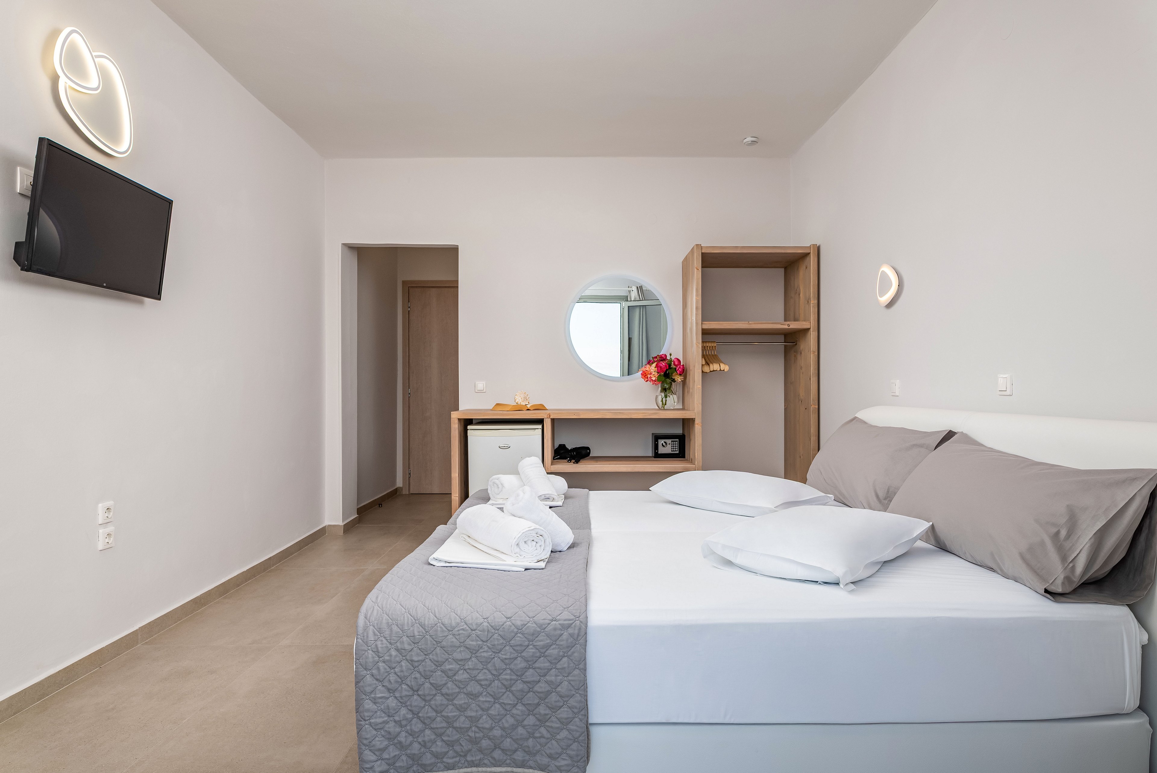 Bright, minimalist hotel bedroom featuring a neatly made double bed with white linens and gray pillows. The room includes a wall-mounted flat-screen TV, open wardrobe, and a small desk with a round mirror and colorful flowers. Soft, modern wall lights add a warm ambiance to the neutral-toned space. Fresh towels are folded on the bed, enhancing the clean and welcoming feel of the room.