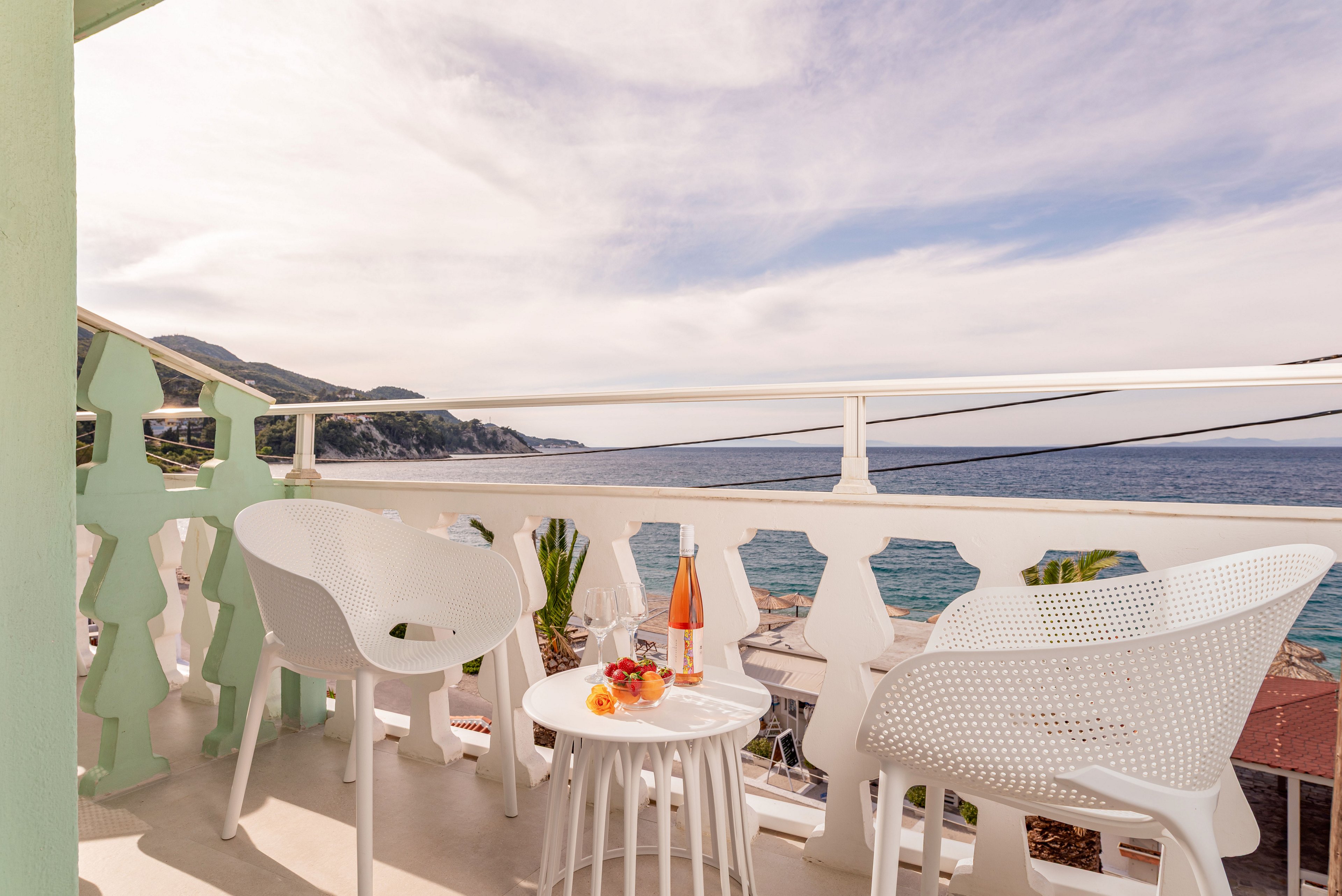 A small seaside balcony features two modern white chairs and a round table set for drinks. A bottle of rosé wine, two wine glasses, and a plate of fruit sit on the table, suggesting a relaxed moment. The balcony railing overlooks a calm blue sea with distant hills under a partly cloudy sky. Soft pastel tones and bright sunlight create a warm, inviting holiday atmosphere.
