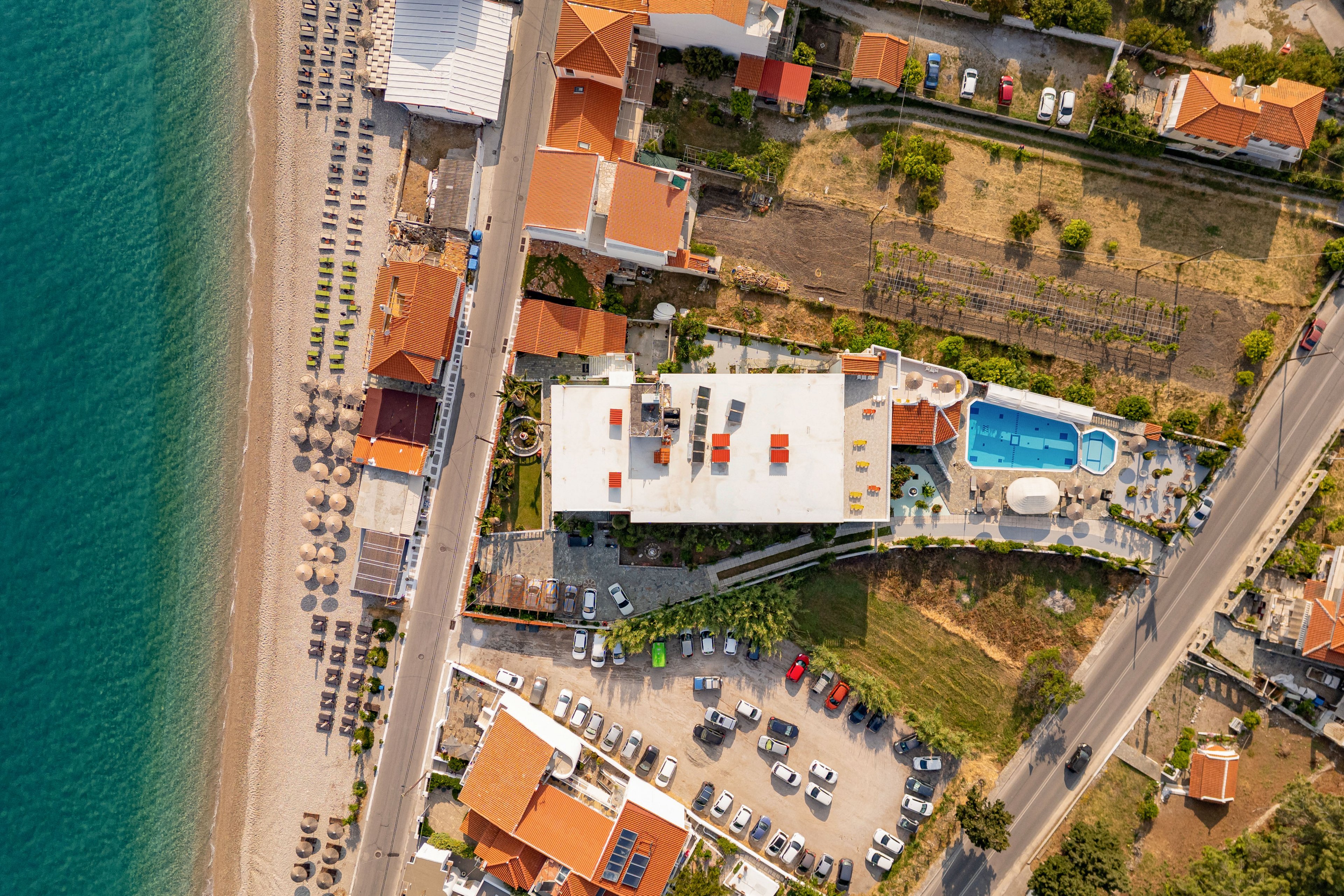A drone-captured aerial view shows a coastal resort complex beside a sandy beach and turquoise sea. The central building has a flat rooftop with seating areas, surrounded by smaller orange-roofed structures and a curved swimming pool. Sun loungers and umbrellas line the beach, while a parking lot filled with cars sits between the resort and the main road. The scene is bathed in warm sunlight, highlighting the orderly layout and relaxed holiday atmosphere.