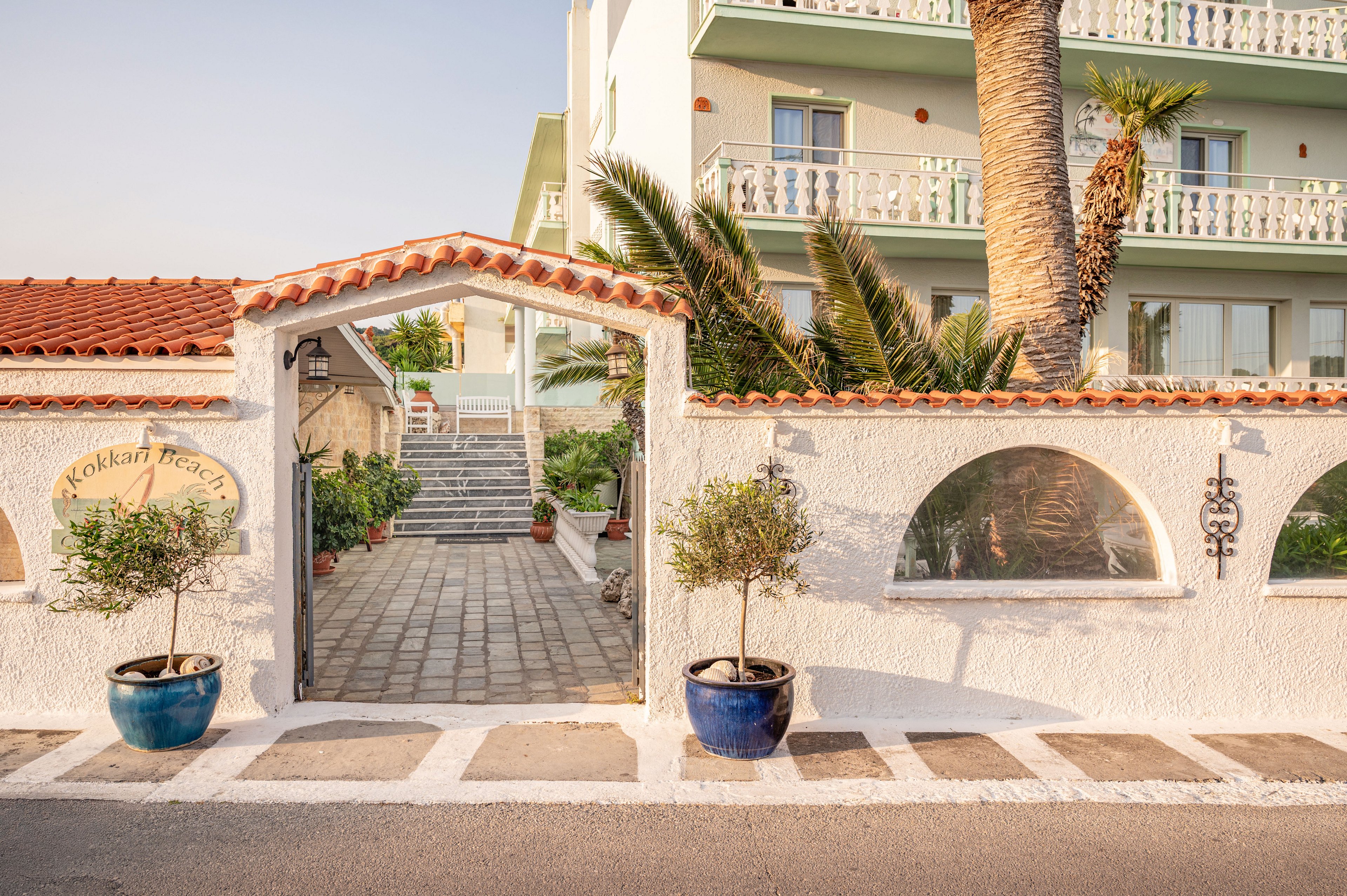Sunlit entrance to a Mediterranean-style hotel features a white stucco wall and tiled archway leading to stone steps. Potted plants in blue and terracotta pots frame the gateway, with palm trees rising behind the wall. The building in the background has balconies with decorative railings and soft pastel colors. Warm evening light enhances the relaxed, coastal atmosphere of the scene.