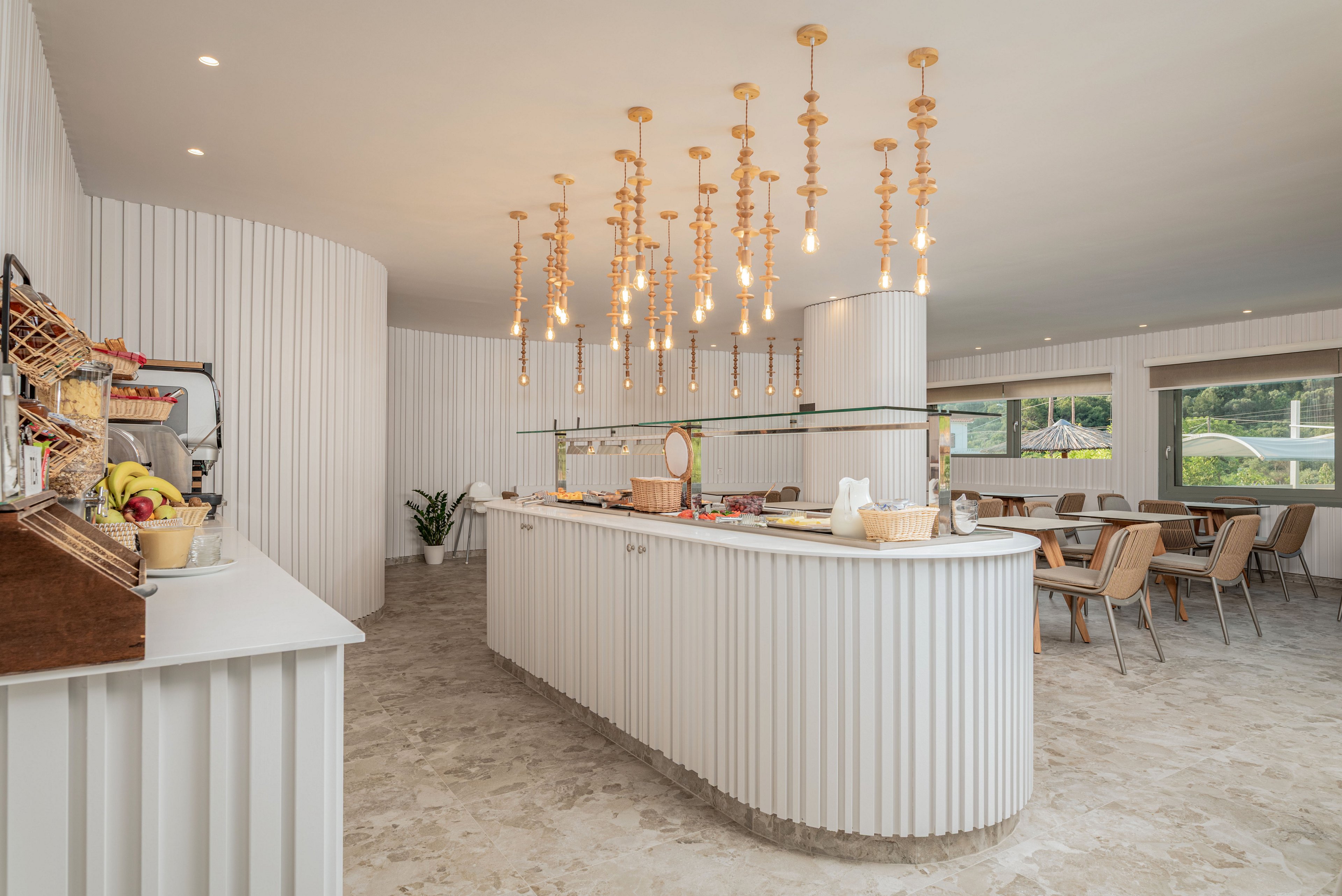 Bright contemporary dining room with a long central buffet counter and hanging pendant lights. The space features light neutral tones, ribbed white wall panels, and stone-textured flooring. Wicker chairs and simple wooden tables create a relaxed café-style seating area near large windows. Fresh fruit and breakfast items are arranged neatly on the counter, suggesting a hotel or resort breakfast setting.