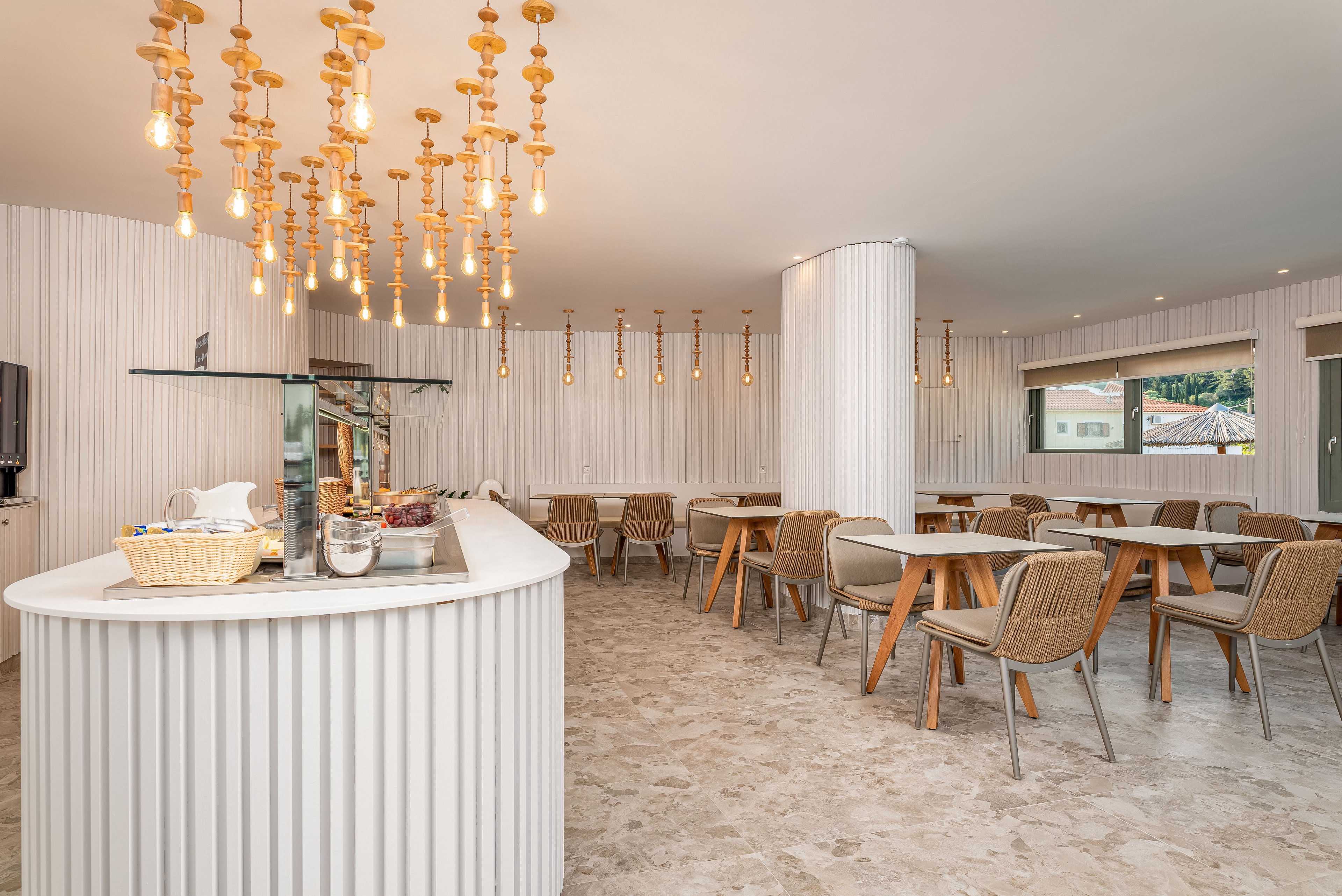 Bright contemporary dining area with a curved white buffet counter in the foreground. Numerous warm pendant bulbs hang from the ceiling, creating a cozy atmosphere. Neutral-toned chairs and round tables are arranged neatly across the room. Large windows and light-colored walls enhance the spacious, airy feel of the interior.