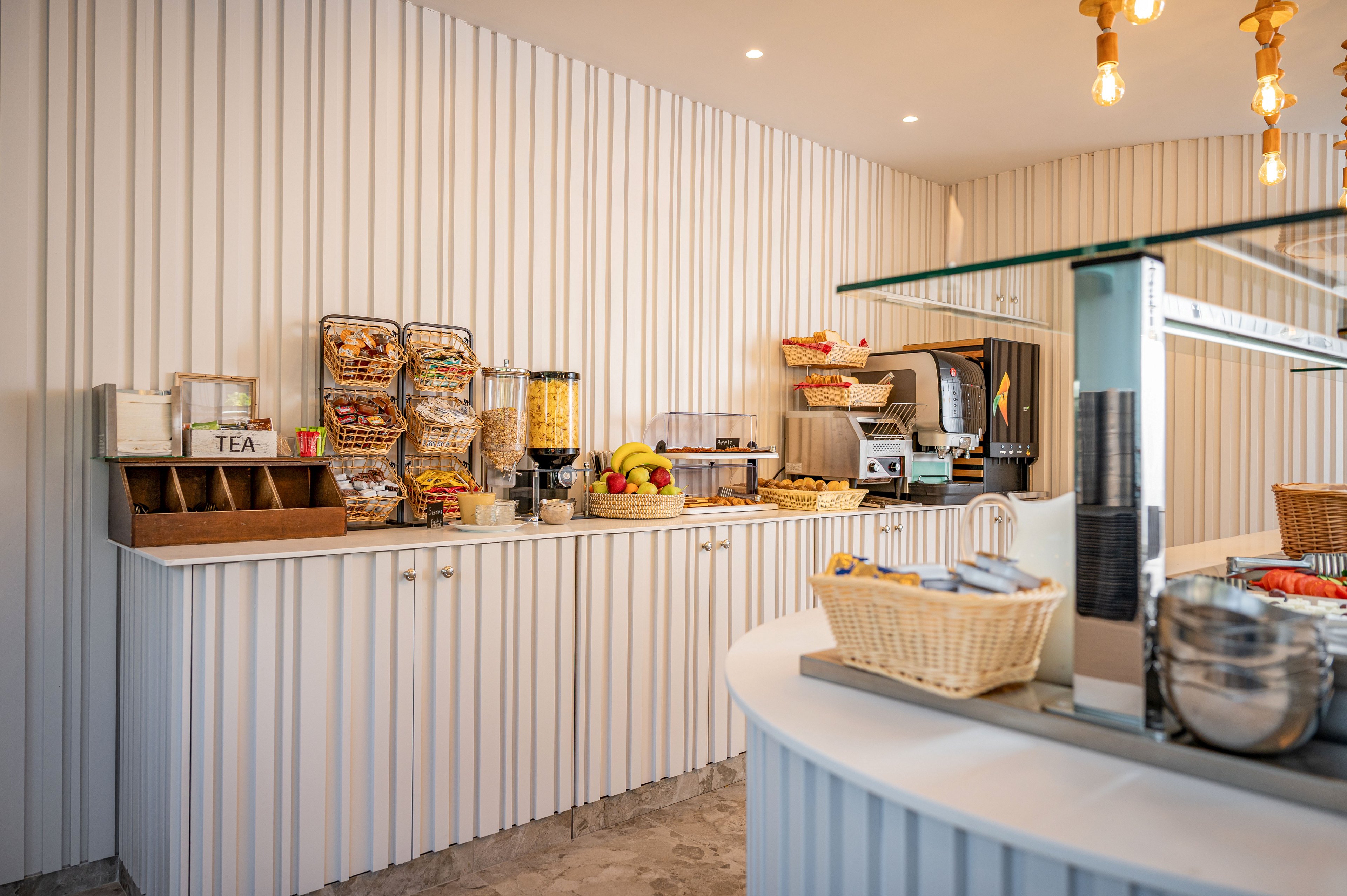 A bright self-service breakfast buffet is set up in a modern hotel dining area. Dispensers with cereals, baskets of bread, and fresh fruit are arranged neatly along a white counter. A toaster, juice machine, and other breakfast appliances are visible in the background. A wooden box labeled TEA holds assorted tea options, while wicker baskets and warm lighting add a cozy touch.