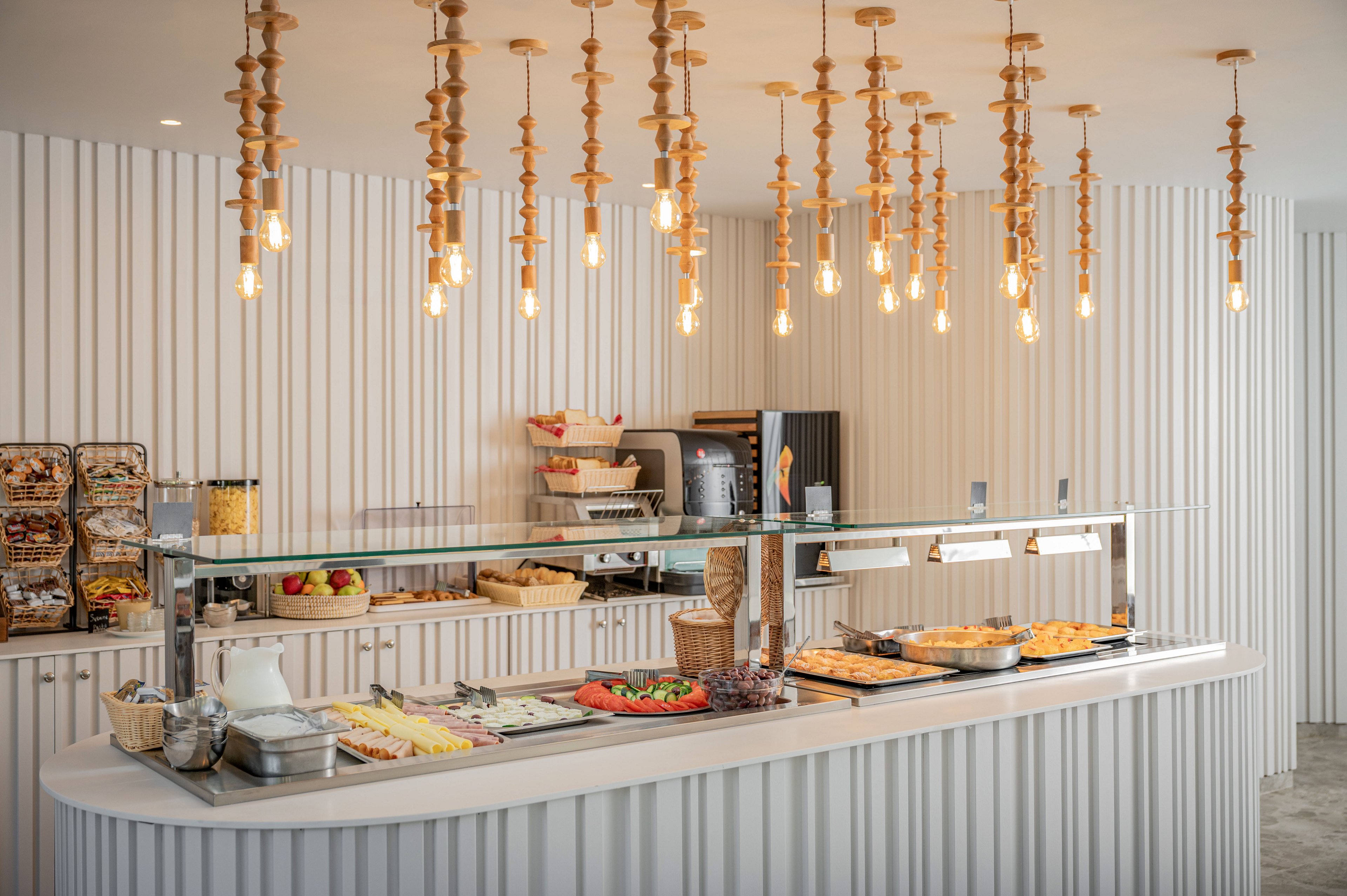 A contemporary self-service buffet counter is set in a minimalist cafeteria with light vertical wall panels. Various fresh foods, including fruits, salads, and baked items, are displayed along the curved counter under glass and warming lamps. Wooden pendant lights with exposed bulbs hang from the ceiling, creating a warm and inviting atmosphere. The overall design is clean, organized, and modern, emphasizing a casual dining experience.