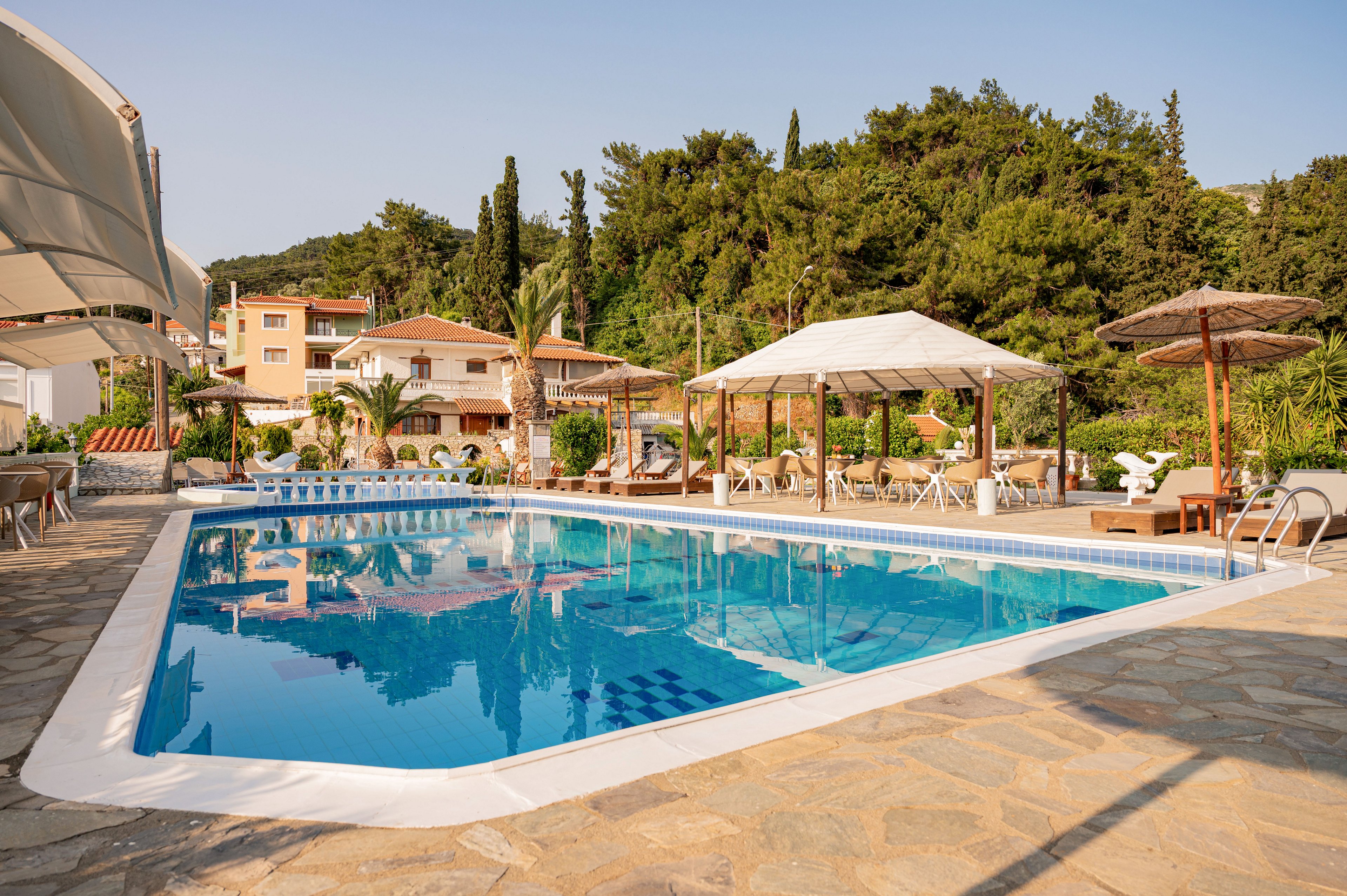Outdoor swimming pool at a holiday resort is surrounded by sun loungers and shaded cabanas. The setting features stone paving, tropical plants, and trees on a hillside in the background. Residential-style buildings with balconies overlook the pool area, creating a relaxed vacation atmosphere. Clear blue water reflects the sky, emphasizing a warm, sunny day.
