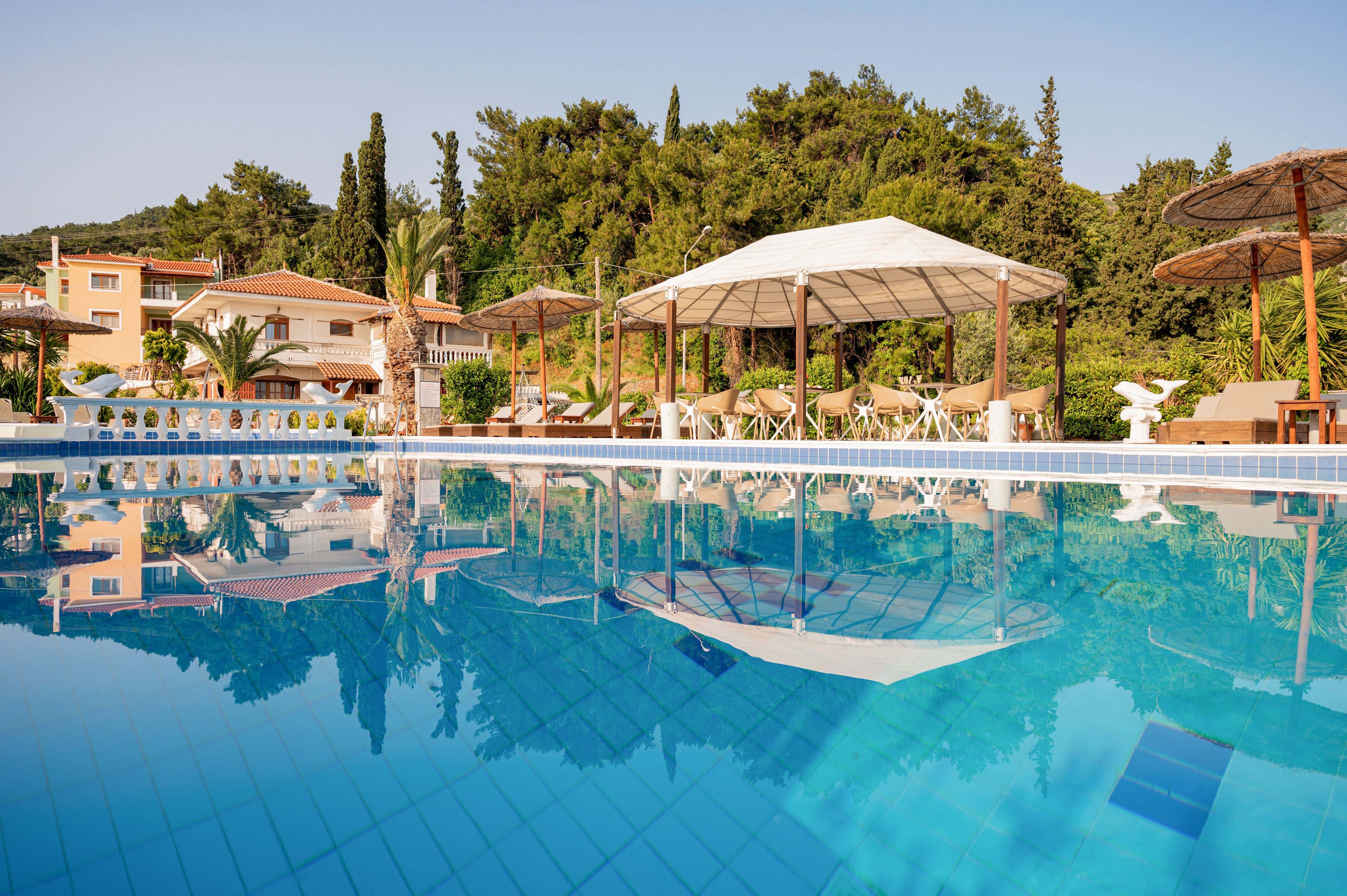 A clear blue swimming pool reflects nearby cabanas and surrounding buildings in a sunny resort setting. White lounge chairs and shaded seating areas line the poolside, creating a relaxed vacation atmosphere. Green trees and hills rise behind the structures, adding a natural backdrop to the leisure scene. The calm water and warm light suggest a peaceful, inviting place for guests to unwind.