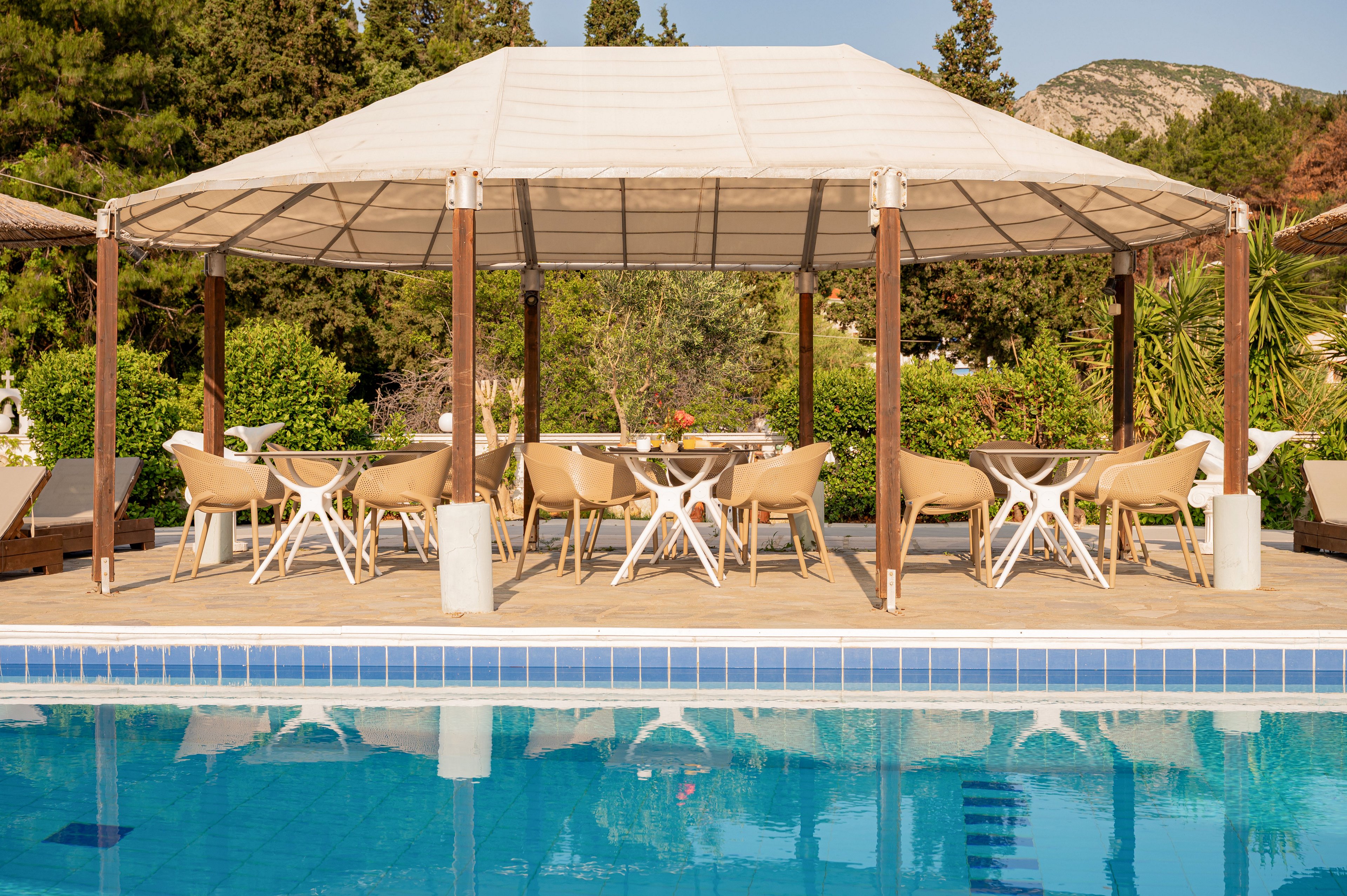 An open-air seating area with modern chairs and tables sits beneath a large canopy beside a clear blue swimming pool. The scene is surrounded by lush green trees and shrubs, with distant hills visible in the background. Sunlight reflects off the calm pool water, creating a bright and inviting vacation atmosphere. The overall setting suggests a relaxed resort or hotel environment ideal for leisure and socializing.