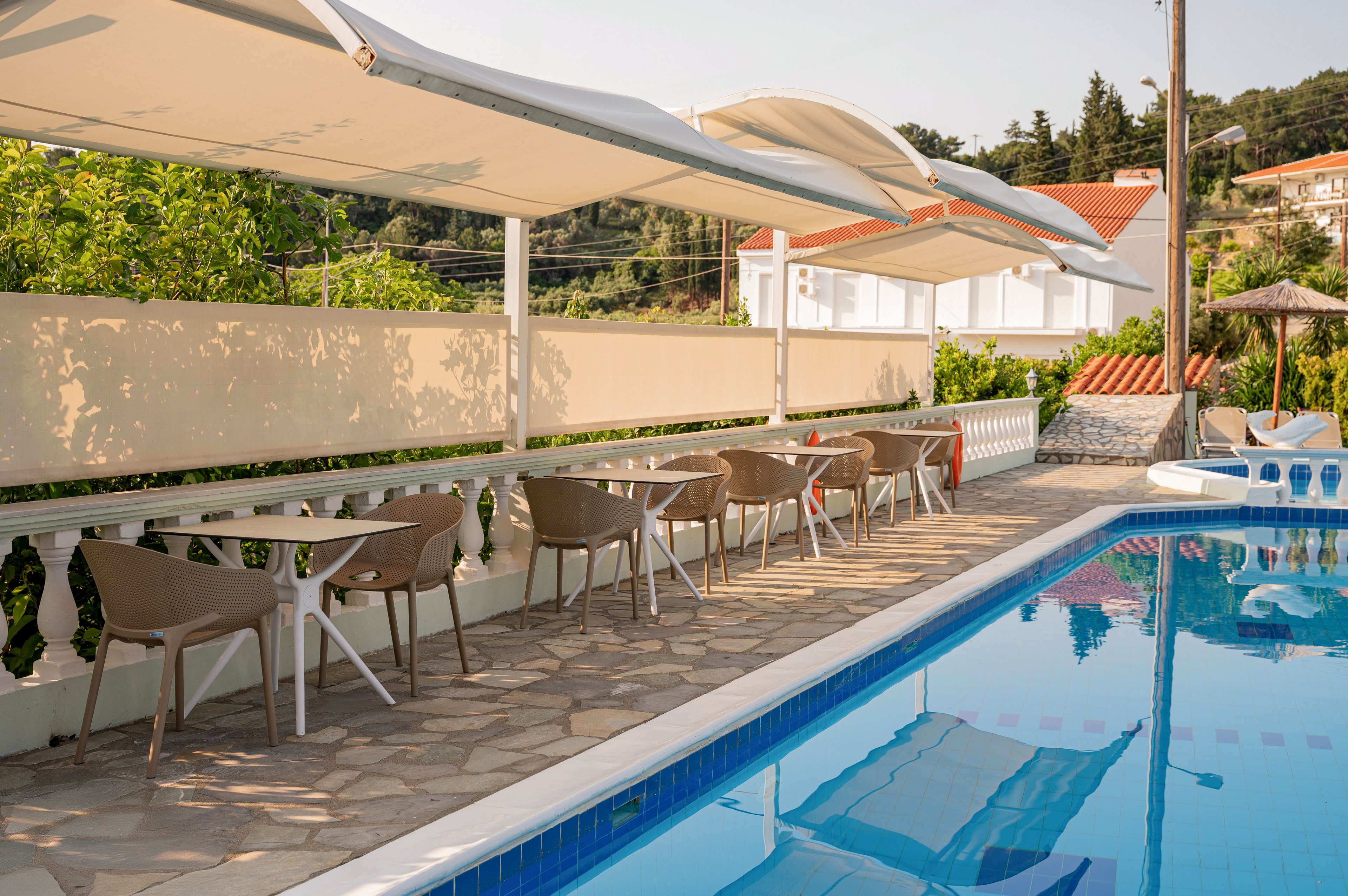 Outdoor resort terrace with modern chairs and small tables lines the edge of a clear blue swimming pool. Large white awnings provide shade above the seating area, while a stone-tiled floor runs alongside the pool. A cream-colored wall separates the terrace from lush green trees and distant houses with red-tiled roofs. The scene is calm and inviting, suggesting a relaxed holiday atmosphere in warm weather.