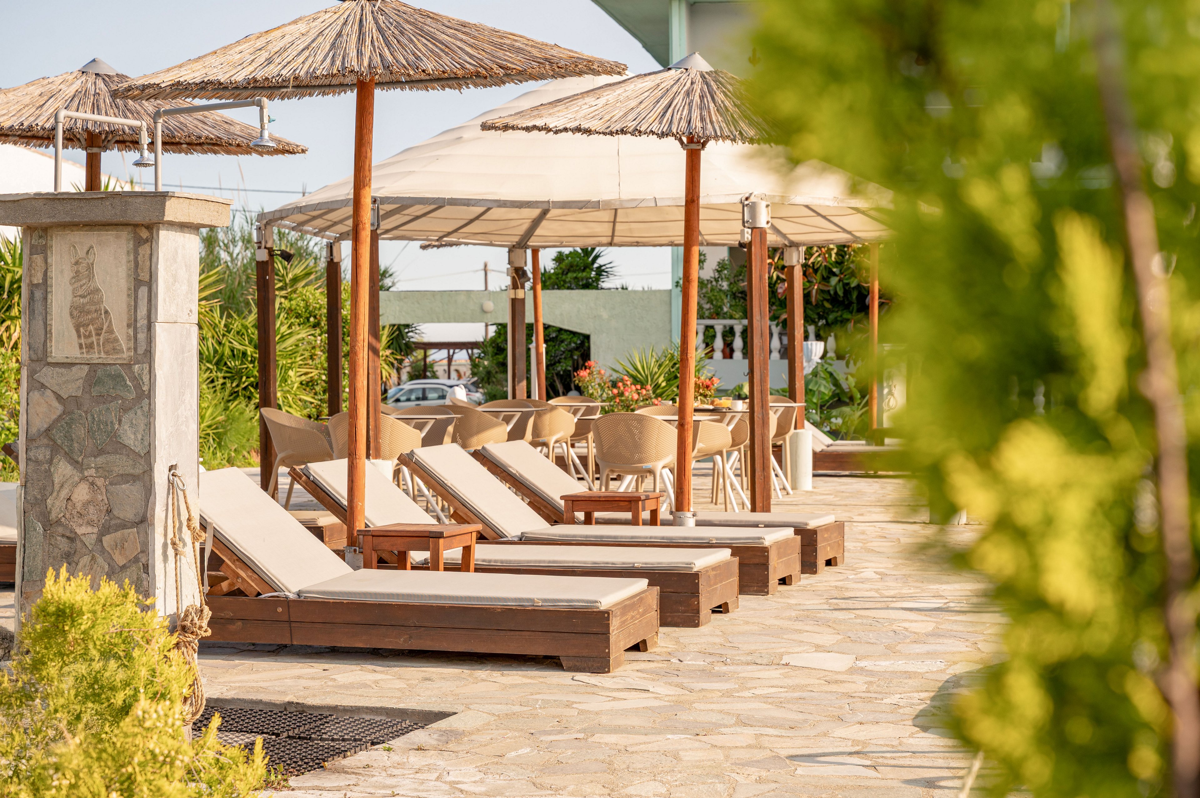 The image shows a row of cushioned wooden sun loungers arranged along a stone-paved terrace. Straw parasols and large white canopies provide shade over the seating area, with wicker chairs and tables visible in the background. Lush greenery and shrubs frame the foreground, adding a natural touch to the resort setting. The warm lighting suggests a sunny, relaxing vacation atmosphere at an outdoor pool or patio area.