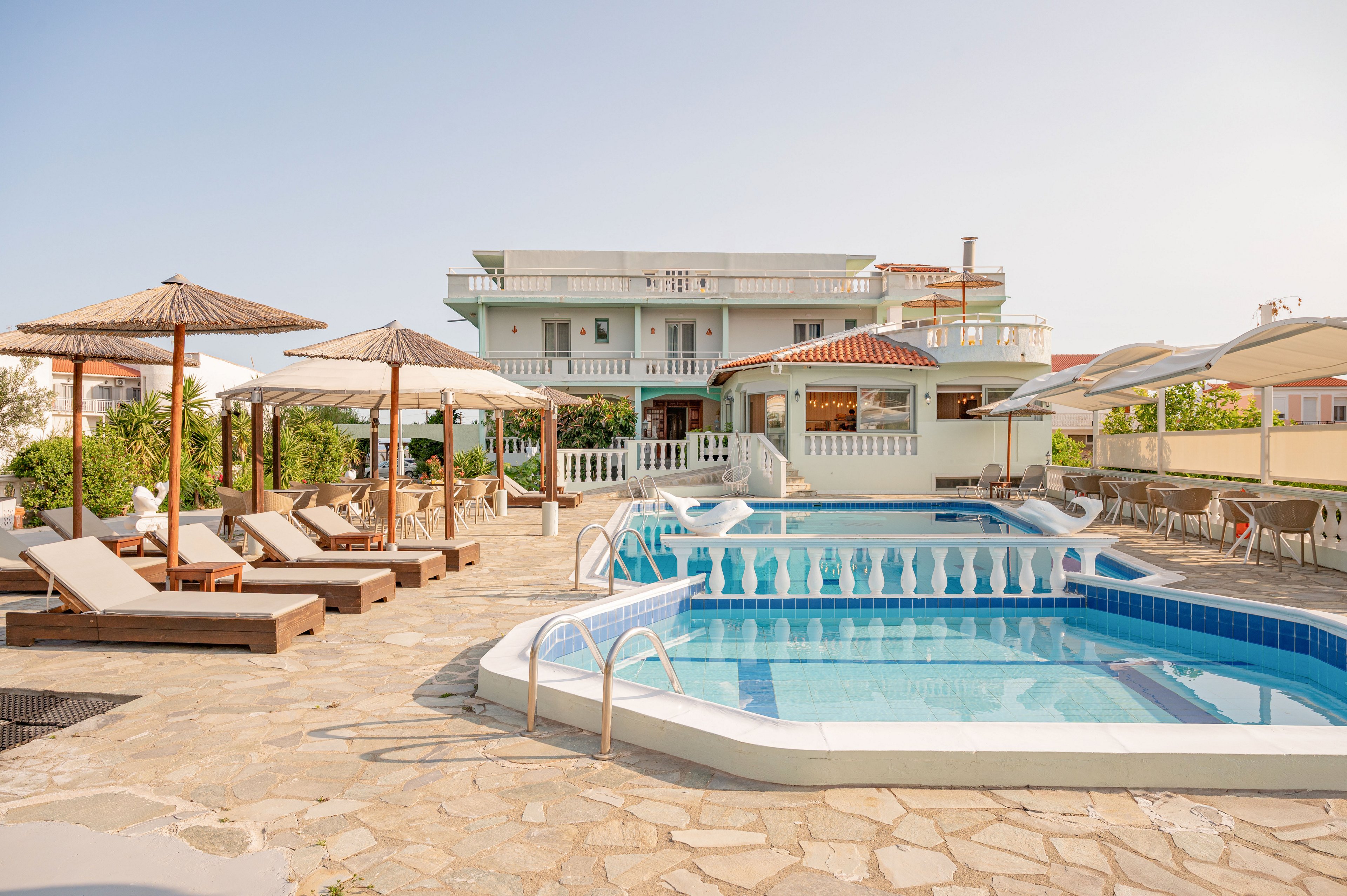 A bright resort courtyard features a clear blue swimming pool surrounded by sun loungers and wooden parasols. The multi-story hotel building in the background has balconies and white railings, creating a Mediterranean-style appearance. Stone paving leads around the pool area, with additional shaded seating and greenery adding to the relaxed atmosphere. The warm daylight and soft colors convey a calm, inviting vacation setting.