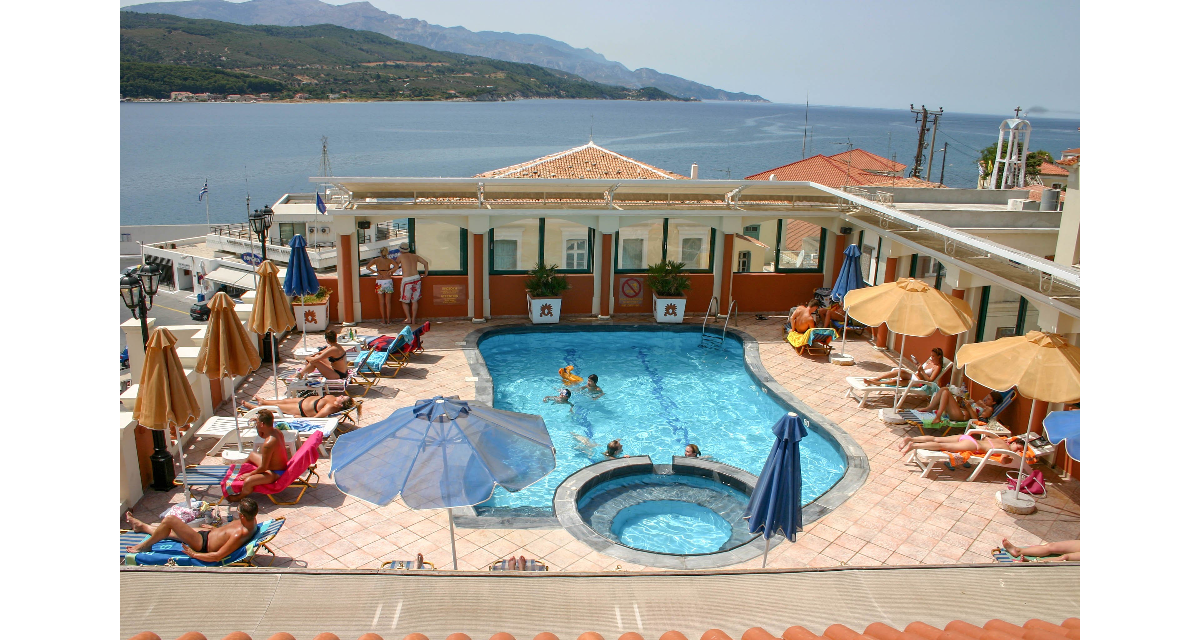 A rooftop hotel swimming pool is surrounded by sun loungers and umbrellas, with guests relaxing and swimming. The pool area includes a smaller circular pool in the foreground, possibly a hot tub or children's pool. Beige tiles and tan umbrellas contrast with the bright blue water and sky. In the background, a calm sea and distant mountains create a scenic coastal view.