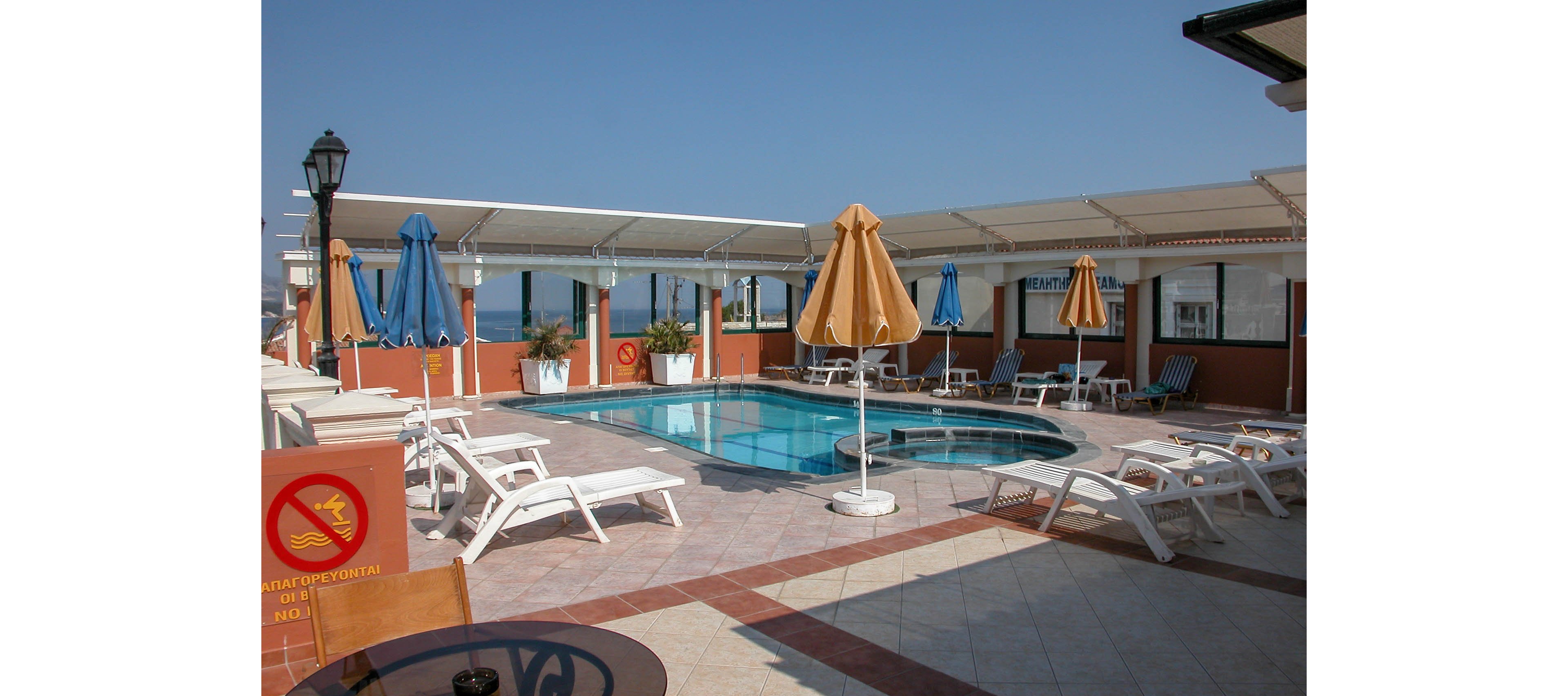 An outdoor rooftop swimming pool is surrounded by white sun loungers and colorful umbrellas under a clear blue sky. The setting appears to be a hotel or resort terrace with tiled flooring and partial sea views in the background. Blue and yellow parasols provide shade around the pool, while a shaded seating area with tables is visible in the foreground. A small sign with a red circle and diagonal line indicates a prohibition near the pool area.