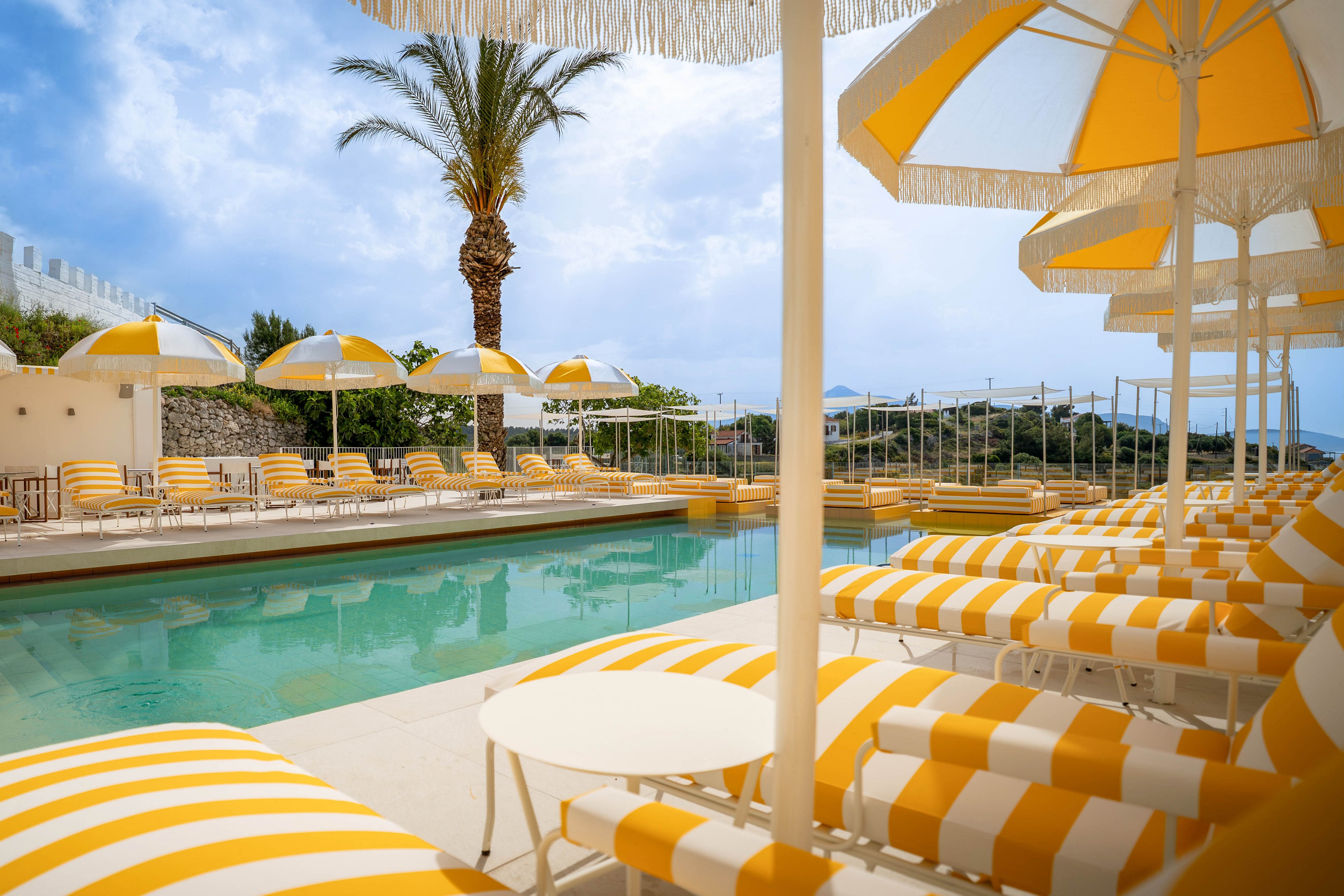 An outdoor resort swimming pool is lined with yellow and white striped sun loungers and umbrellas. The scene is set on a bright day with partly cloudy skies and a calm, inviting atmosphere. A tall palm tree rises behind the pool area, and distant hills and buildings are visible in the background. The overall palette is warm and summery, emphasizing relaxation and vacation vibes.