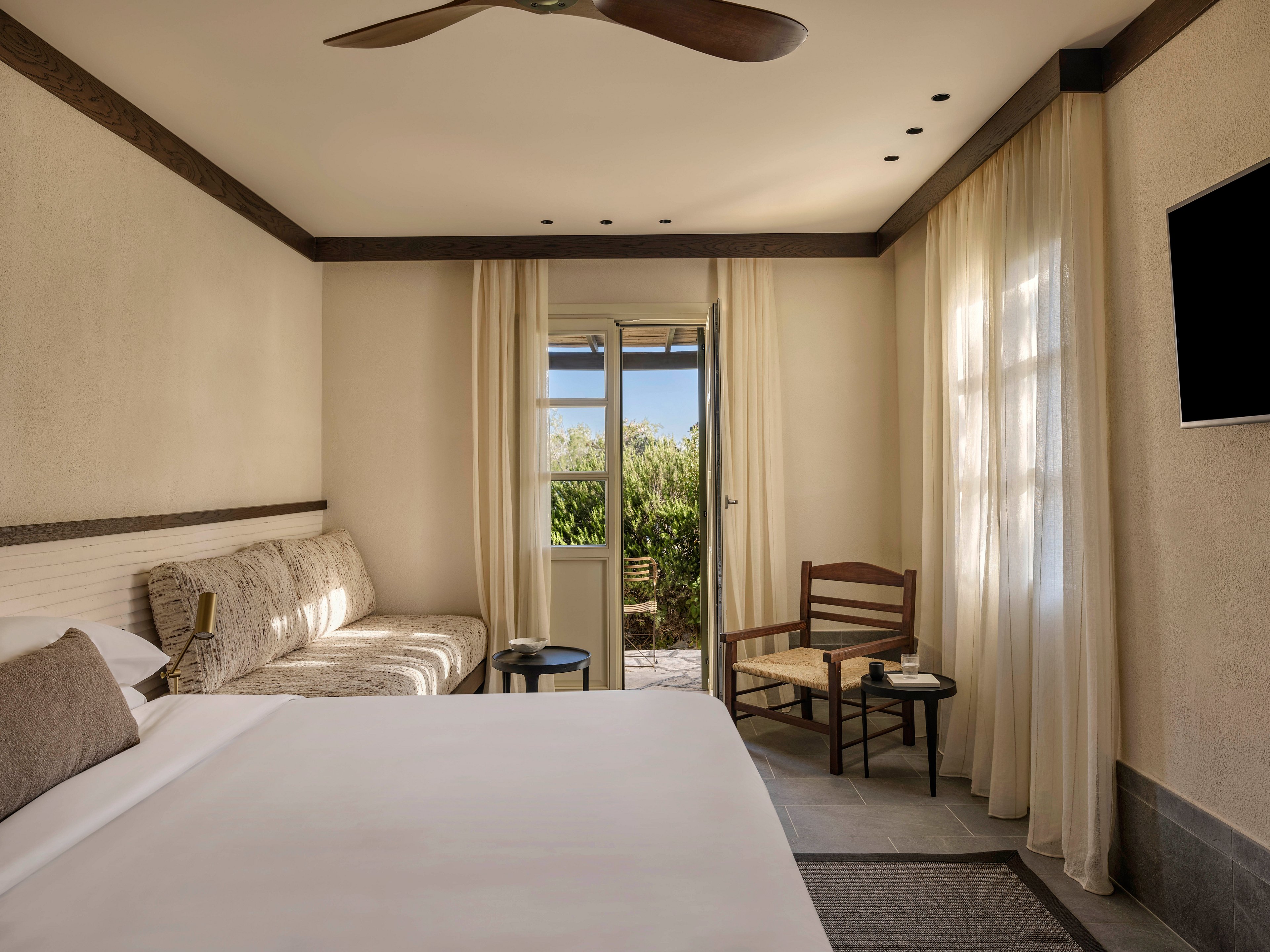 A contemporary hotel room features a large neatly made bed and a cushioned sofa along the wall. Neutral beige walls and light curtains create a calm, minimalist atmosphere. A wooden chair with a small side table sits near the windowed door leading to an outdoor terrace with greenery. Recessed ceiling lights and a ceiling fan add modern comfort, while a wall-mounted television completes the cozy space.