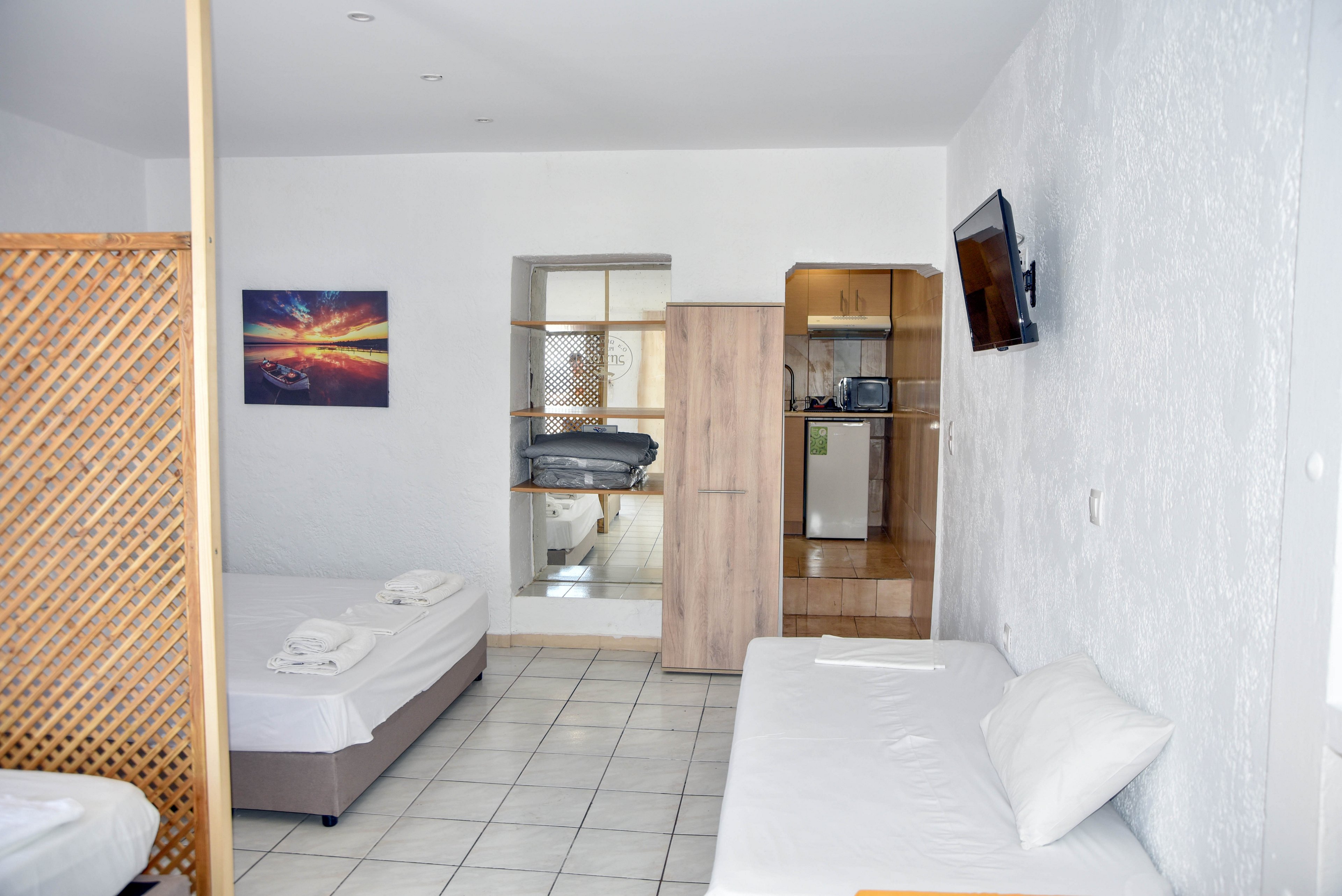A bright, minimalist hotel room features a double bed and a single daybed with white linens. The space includes tiled floors, white walls, and a wooden wardrobe separating the sleeping area from a small kitchenette. A wall-mounted television faces the beds, and a colorful landscape artwork adds contrast to the neutral decor. The room appears clean, modern, and designed for practical, comfortable short stays.