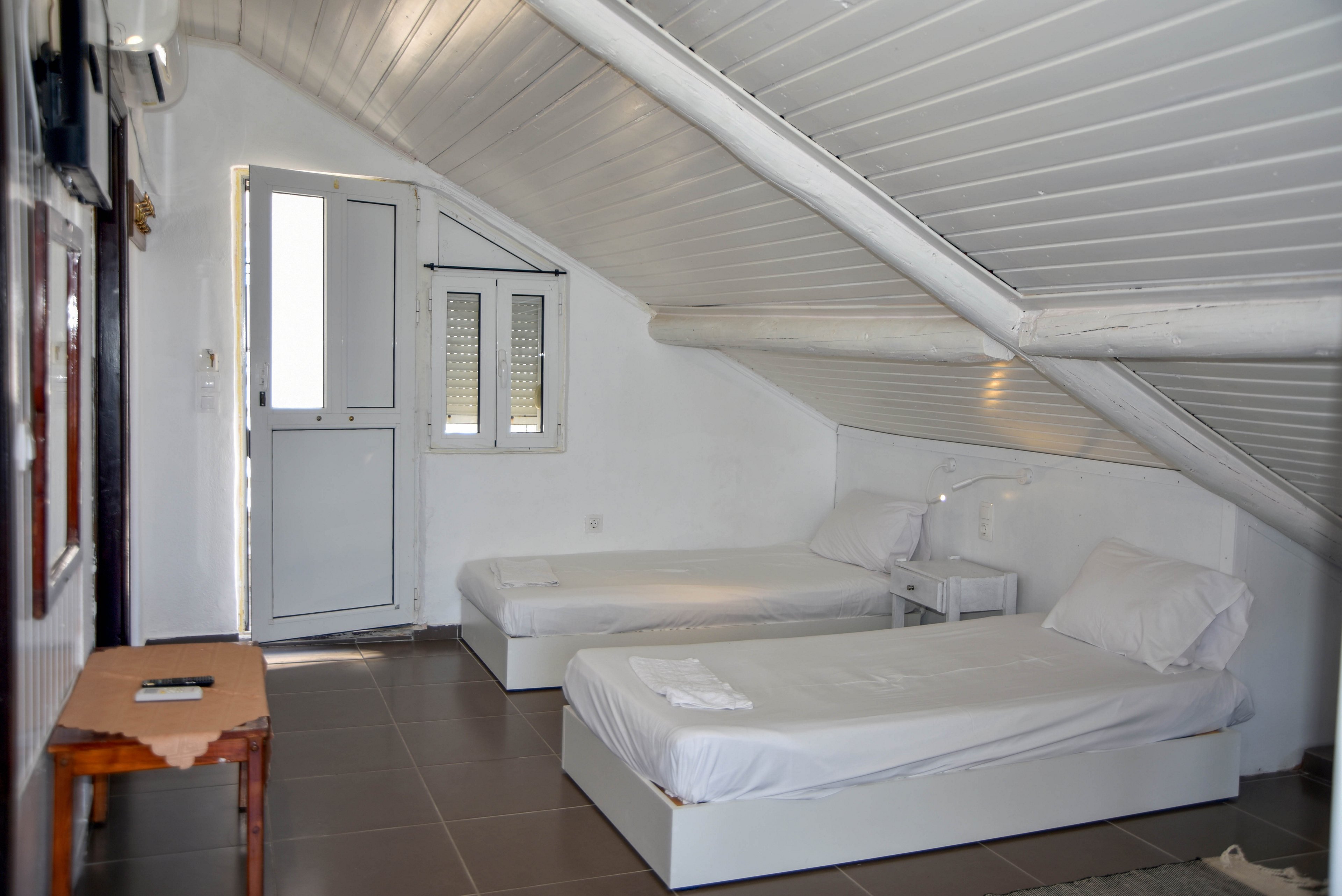 Bright attic bedroom featuring two simple white single beds against a sloped ceiling. The room is painted white with a matching white door and small window letting in natural light. A small bedside table with a lamp sits between the beds, and a wooden table stands near the entrance. The overall decor is minimal and clean, creating a calm and airy atmosphere.