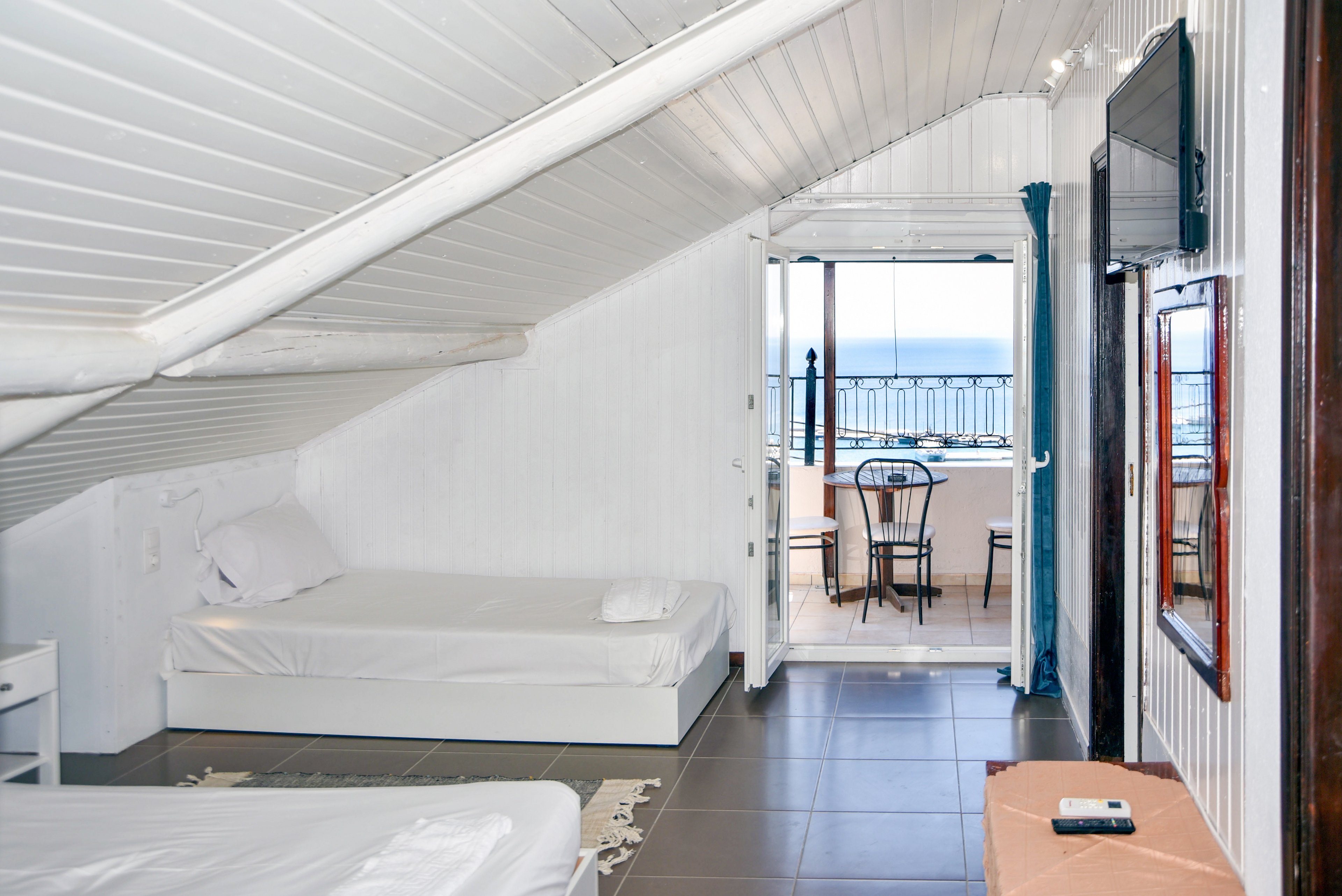 An attic-style hotel room features two simple single beds under a sloped white ceiling. The interior is bright with white walls, dark floor tiles, and minimal furnishings including a wall-mounted television and a small table. Through open doors, a balcony with a small table and chairs overlooks the ocean and a metal railing. Natural light from the balcony enhances the airy, coastal atmosphere of the room.