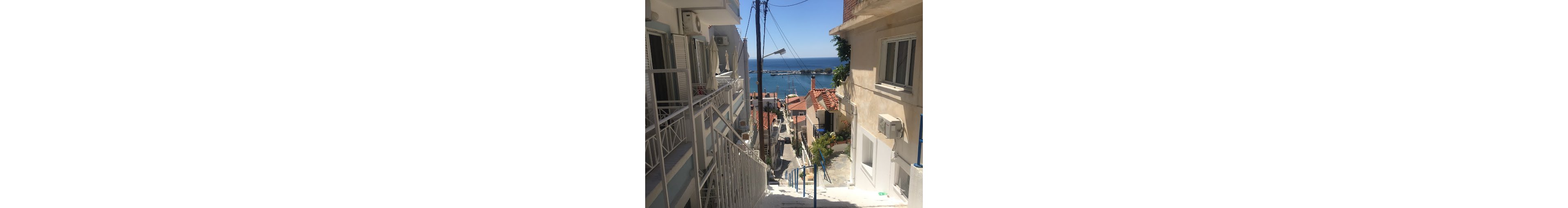 A narrow stairway descends between whitewashed residential buildings toward a bright blue sea. Red-tiled rooftops and balconies line the steep alley, leading the eye down to a small harbor and boats in the distance. Sunlight casts strong shadows on the steps and walls, emphasizing the Mediterranean architecture. The scene feels quiet and inviting, suggesting a peaceful seaside town on a clear summer day.