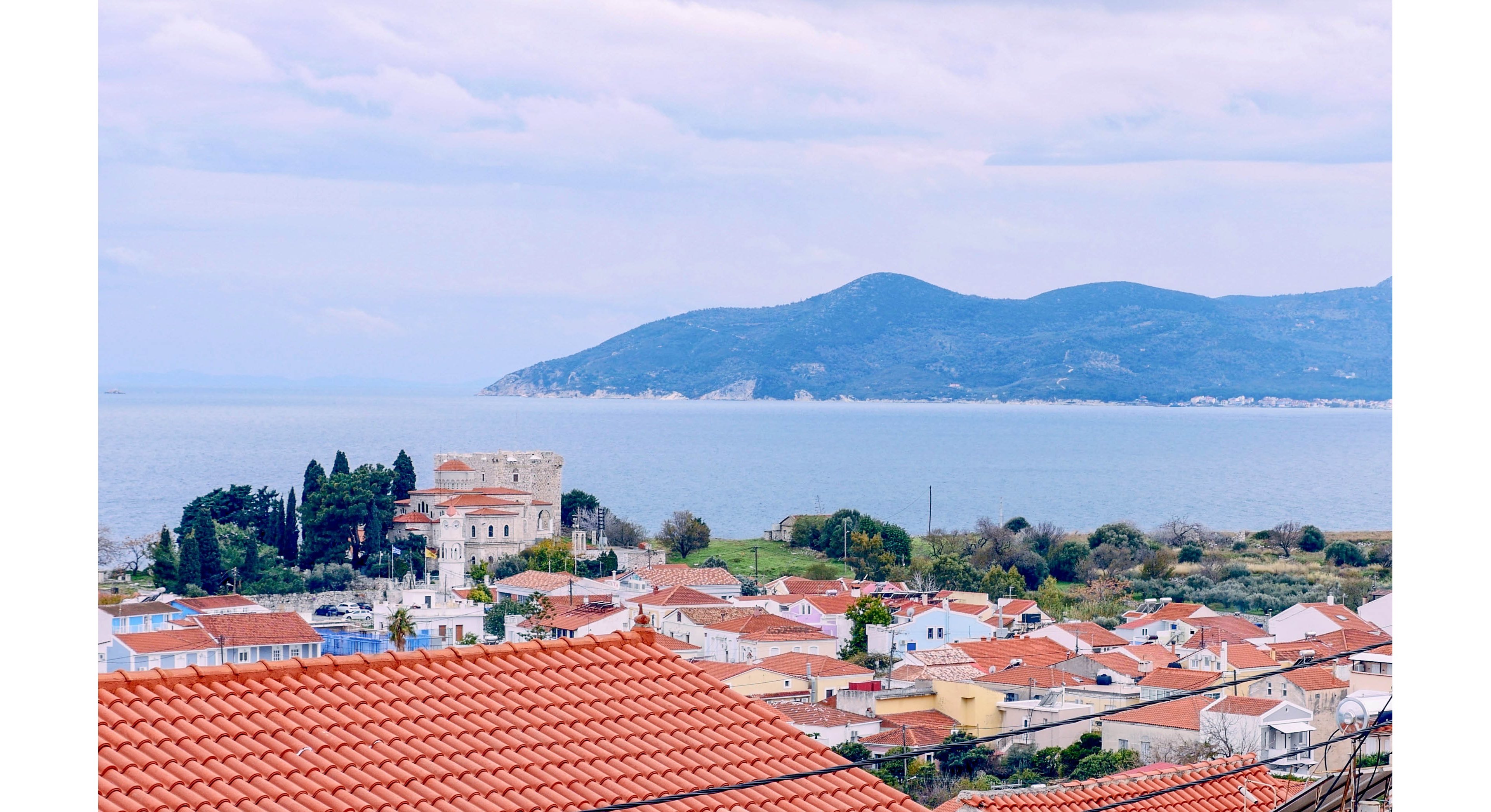 A coastal village with terracotta-tiled rooftops stretches toward a calm blue bay. In the midground, a light-colored stone building and clusters of trees stand near the shoreline. Rolling green hills and a mountainous peninsula rise across the water under a cloudy sky. The scene feels peaceful and picturesque, highlighting Mediterranean-style architecture and natural coastal beauty.