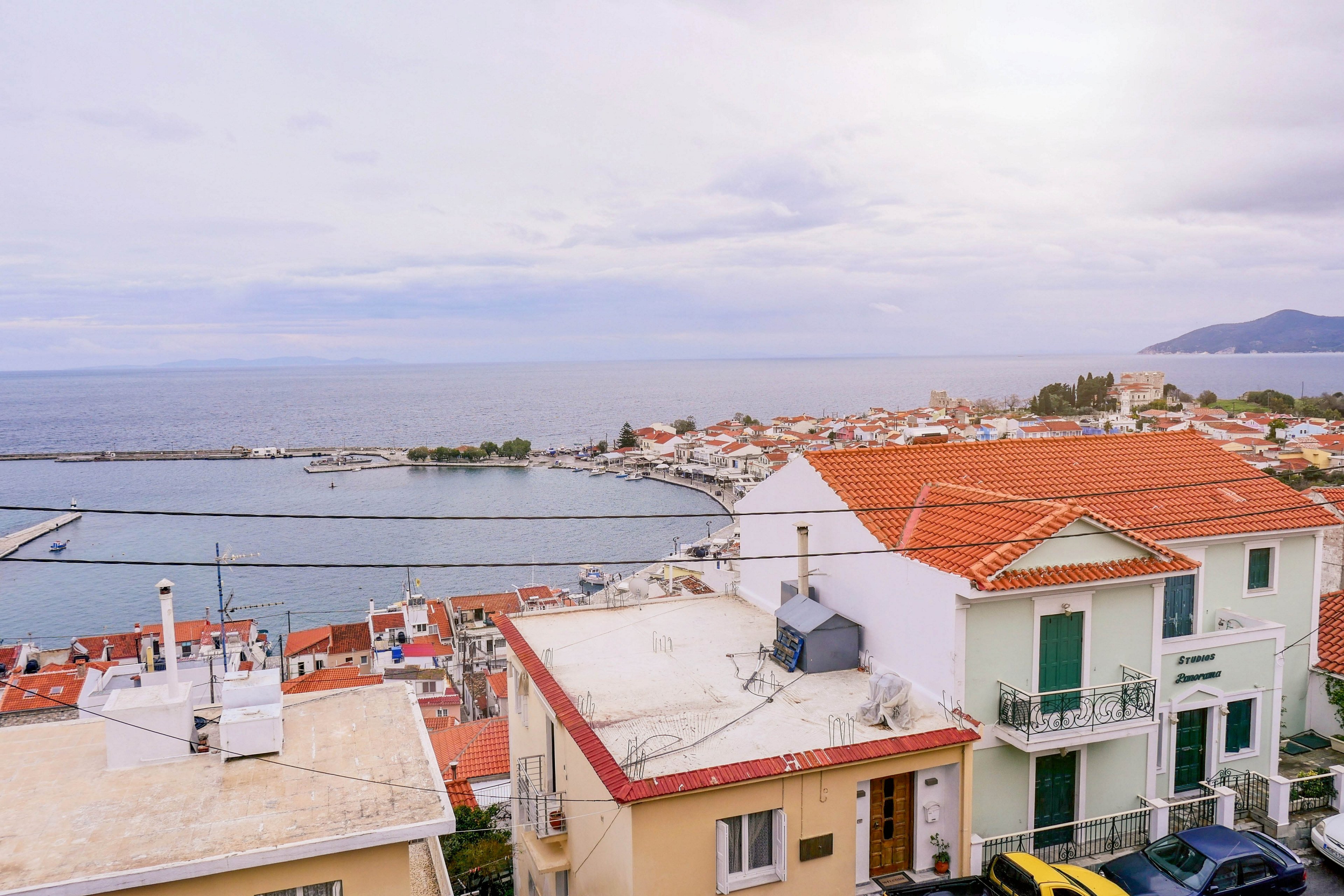 A coastal town with red-tiled rooftops overlooks a calm sea and harbor. Residential buildings with balconies and terraces fill the foreground, leading down toward the waterfront. The curved shoreline and distant breakwater frame the bay under a cloudy sky. Soft light and muted colors create a peaceful, relaxed atmosphere.
