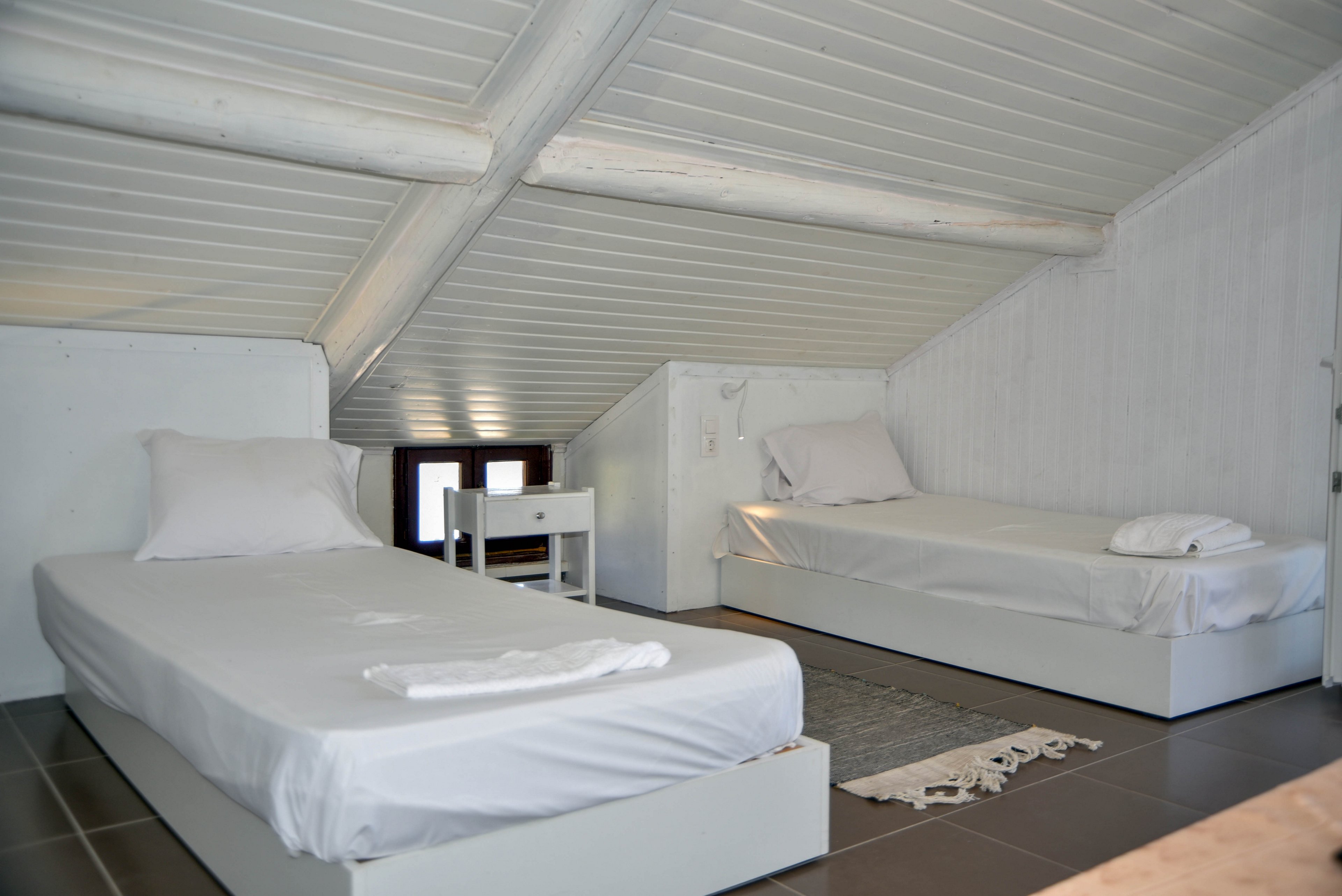 A bright attic bedroom features two simple twin beds with white linens. The room has a low, sloped ceiling with exposed white beams and a small window at the far end. A compact white nightstand sits between the beds, and a neutral rug softens the tiled floor. The overall decor is minimalist, emphasizing clean lines and a calm, airy atmosphere.